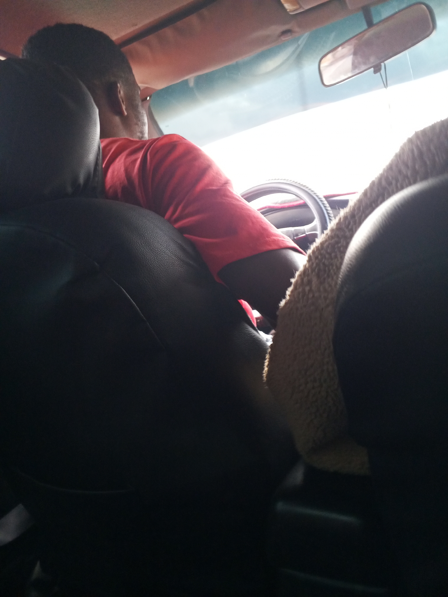 The image shows the interior of a vehicle, likely a car, in Bukuru, Nigeria. A person, viewed from the back and side, is seated in the driver's seat, wearing a red shirt. The person's head and upper torso are visible. The steering wheel of the vehicle is in the foreground, and a fuzzy brown seat cover is draped over a portion of it. The vehicle's interior includes black leather seats, a rear-view mirror, and the roof lining of the car. The scene is illuminated by natural light coming from outside, suggesting it is daytime. The overall impression is a candid shot taken from the back seat of a car.