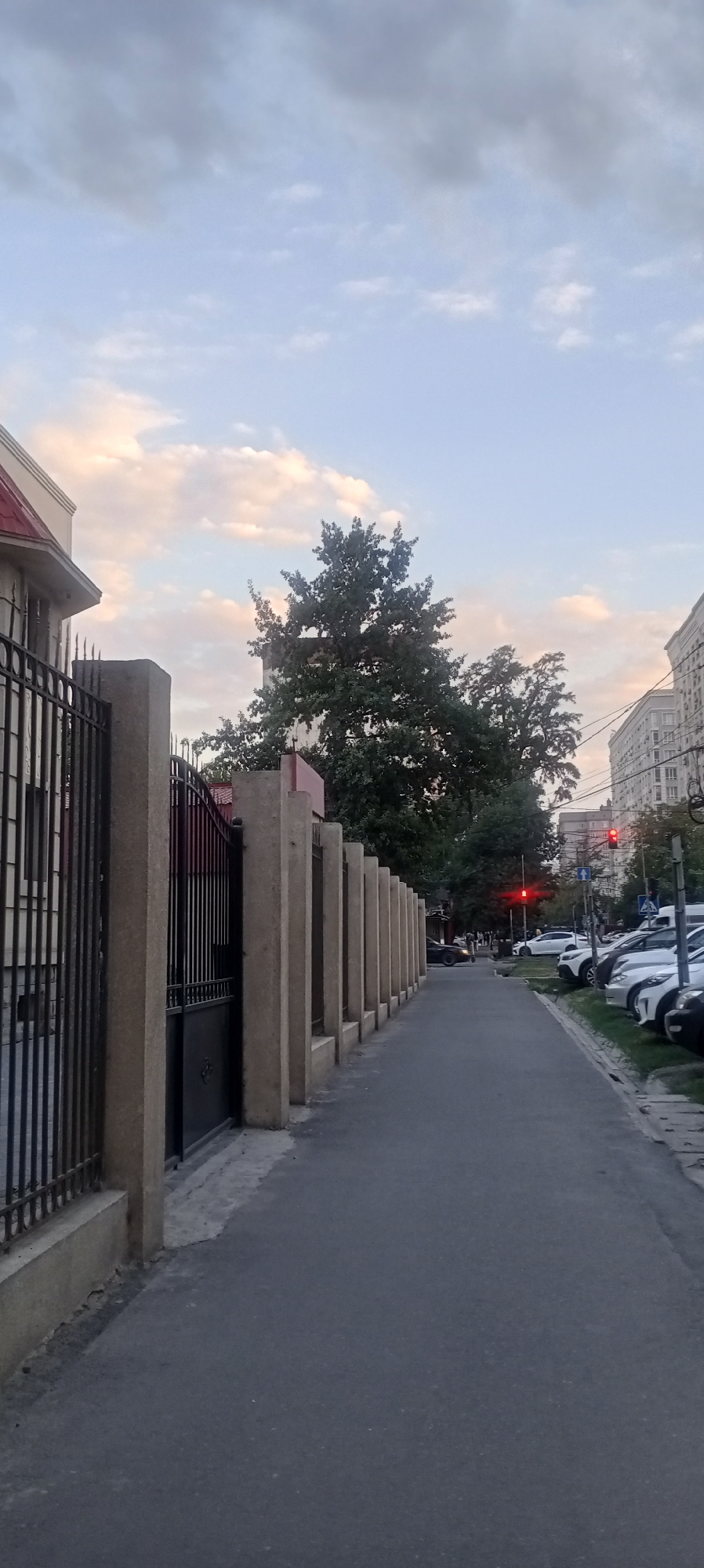 The image depicts a street scene in Bishkek, Kyrgyzstan. A tall black metal fence with concrete pillars runs along the left side of a paved sidewalk. Beyond the fence, a building with a red roof is partially visible, along with a large green tree. The sidewalk extends into the distance, where parked cars and a traffic light are present. The sky is partly cloudy, with a mix of light and dark clouds. The overall impression is that of a quiet urban street, likely during the daytime.