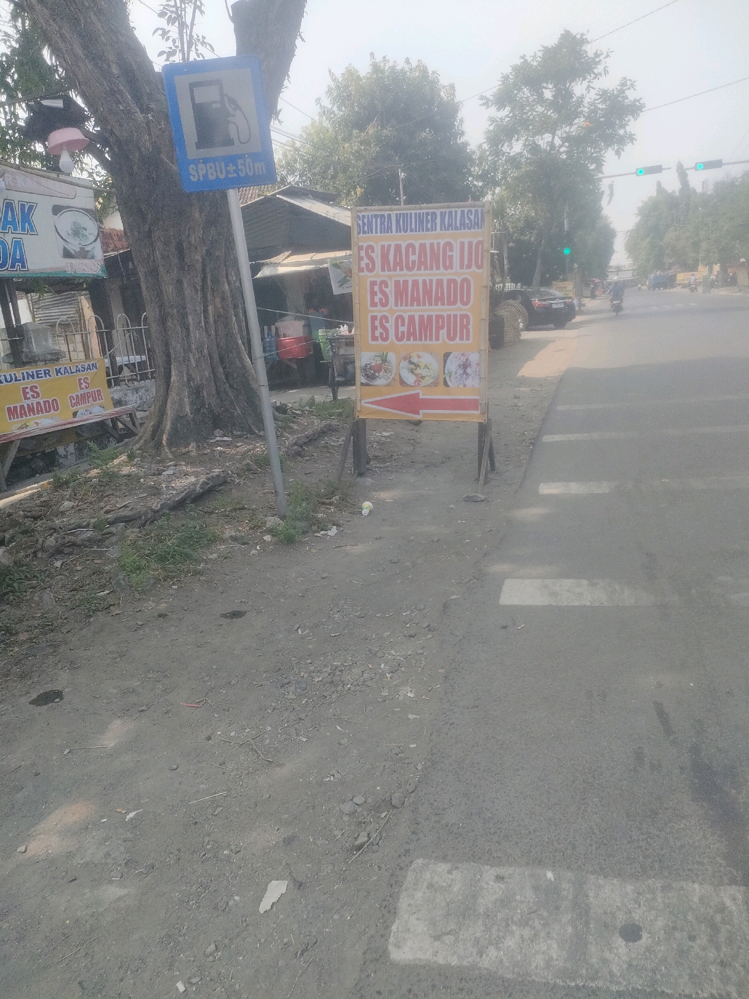 The image shows a street scene in Surabaya, Indonesia. On the left, a large tree trunk stands near a road. A blue sign indicating a gas station (SPBU) with "±50m" is attached to the tree. In front of the tree, there is a yellow sign advertising "SENTRA KULINER KALASAN ES KACANG IJO ES MANADO ES CAMPUR" with pictures of food items. Further to the left, a white sign with blue text reads "DA".
In the background, there are trees, buildings, a traffic light showing green, and a few vehicles including a car and motorcycles. The road has a pedestrian crossing marked with white stripes. The ground is a mix of dirt and gravel. The overall lighting suggests it is daytime, possibly with some haze.