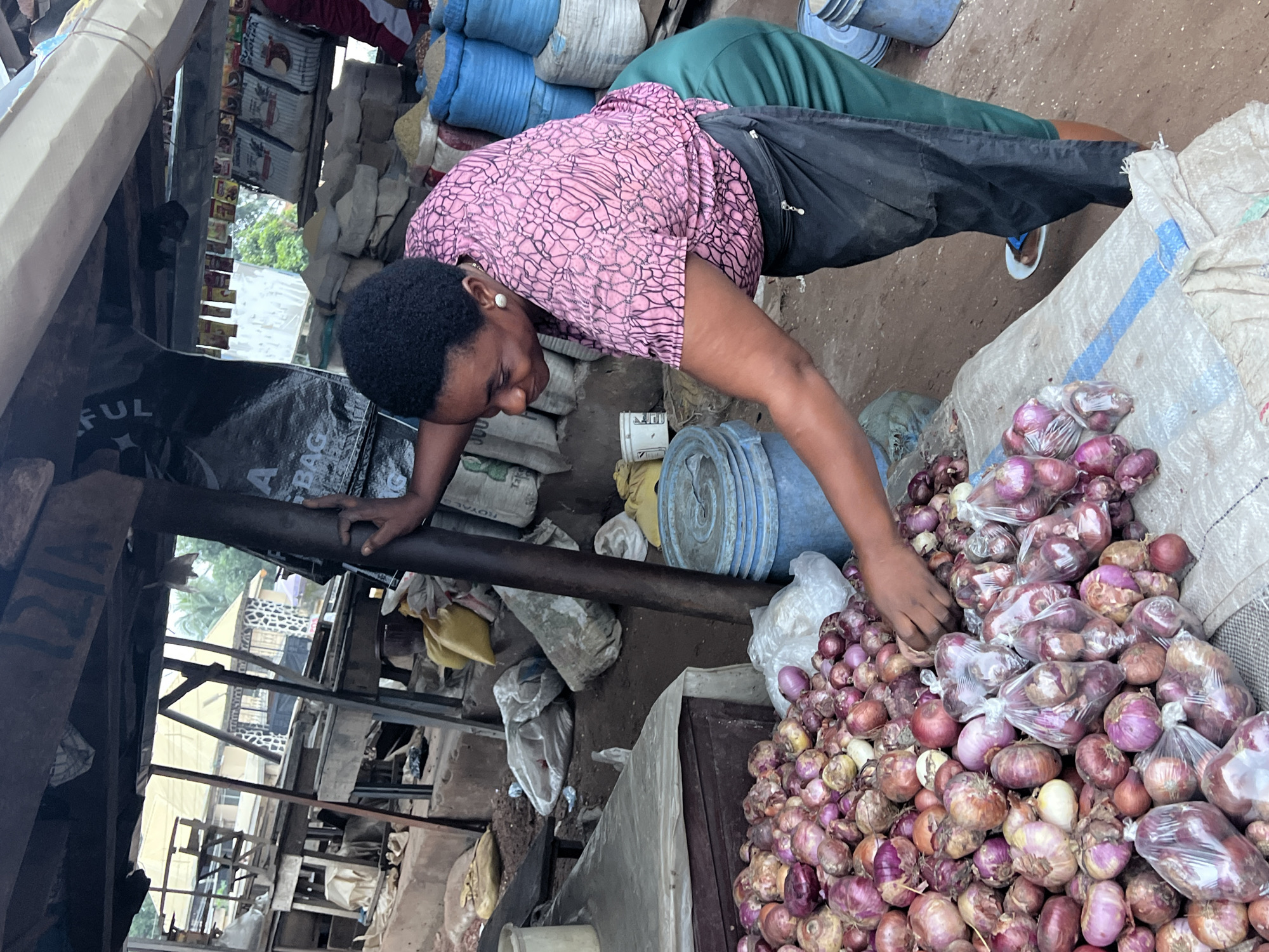 In Nkpor, Nigeria, a woman with dark skin and short, black hair is standing at an outdoor market stall. She is wearing a pink blouse with a white, intricate pattern and a black apron or skirt. Her right arm is extended, and her hand is resting on a dark pole supporting the market's overhead structure. Her gaze is directed downwards towards a large pile of red onions spread across a textured, light-colored surface, possibly a tarp or burlap sack. Some of the onions are packaged in clear plastic bags. Behind her, stacks of blue plastic buckets are visible. The market appears to be a busy environment with various goods, including bags of grains and other produce, displayed in the background. The ground is dirt, and there are wooden structures and stalls typical of an open-air market.