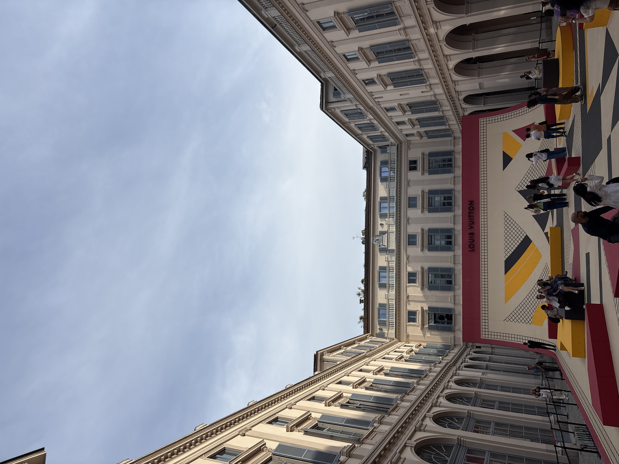 Captured from a low-angle perspective, the image features a view of the sky between two buildings, framed by a large Louis Vuitton display. The building architecture is characterized by multiple windows with light blue shutters and an arched design along the bottom floors. The Louis Vuitton display is situated on the ground level, with the brand's name prominently displayed, and features a geometric pattern in yellow, black, and white. Several people stand around the display, observing and interacting with the exhibit. The sky is light blue with some wispy clouds. The setting is the Piazza del Duomo in Milan, Italy, as identifiable by the architectural style and brand exhibition.