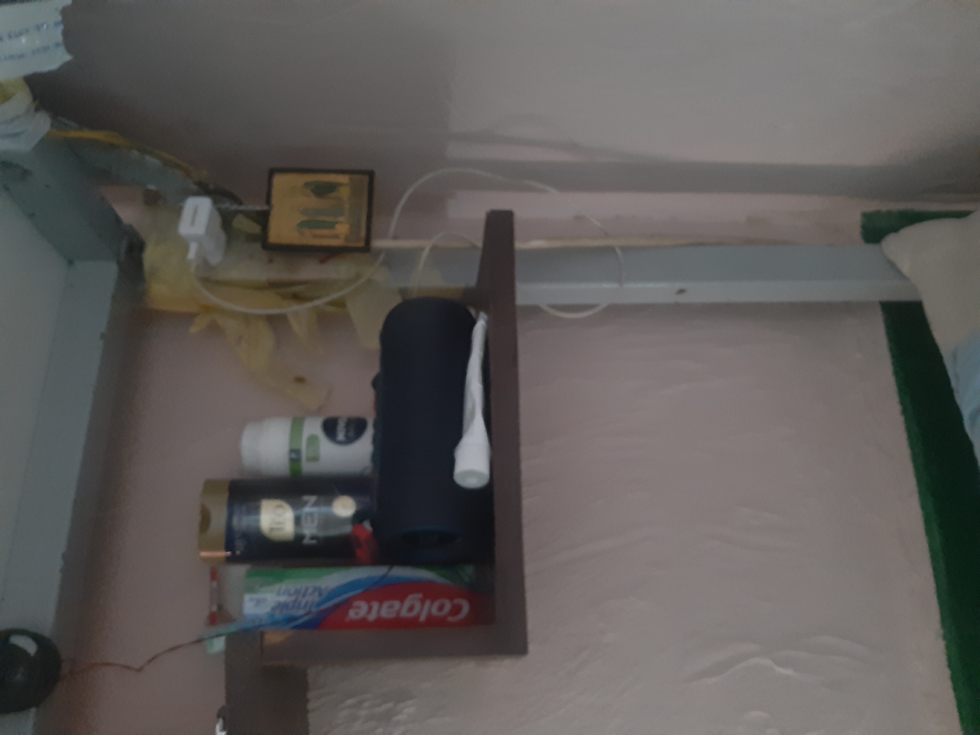 In the city of Cricova, Moldova, an image captures a close-up view of a shelf unit against a textured, light-colored wall. On the shelf, several personal care items are visible. To the left is a tube of Colgate toothpaste with its distinctive red and white packaging. Next to it, a dark cylindrical bottle labeled "Teo MEN" stands upright. Adjacent to this is a white bottle with a green label, possibly another toiletry item. A dark, rectangular object, possibly a speaker, is placed in front of these bottles. A white tube, likely a cosmetic or medical product, lies horizontally in front of the dark object. On the right side of the shelf, a small framed icon with figures is displayed. The shelf unit itself appears to be made of wood with a brown finish. The overall environment suggests a bathroom or a personal storage area.