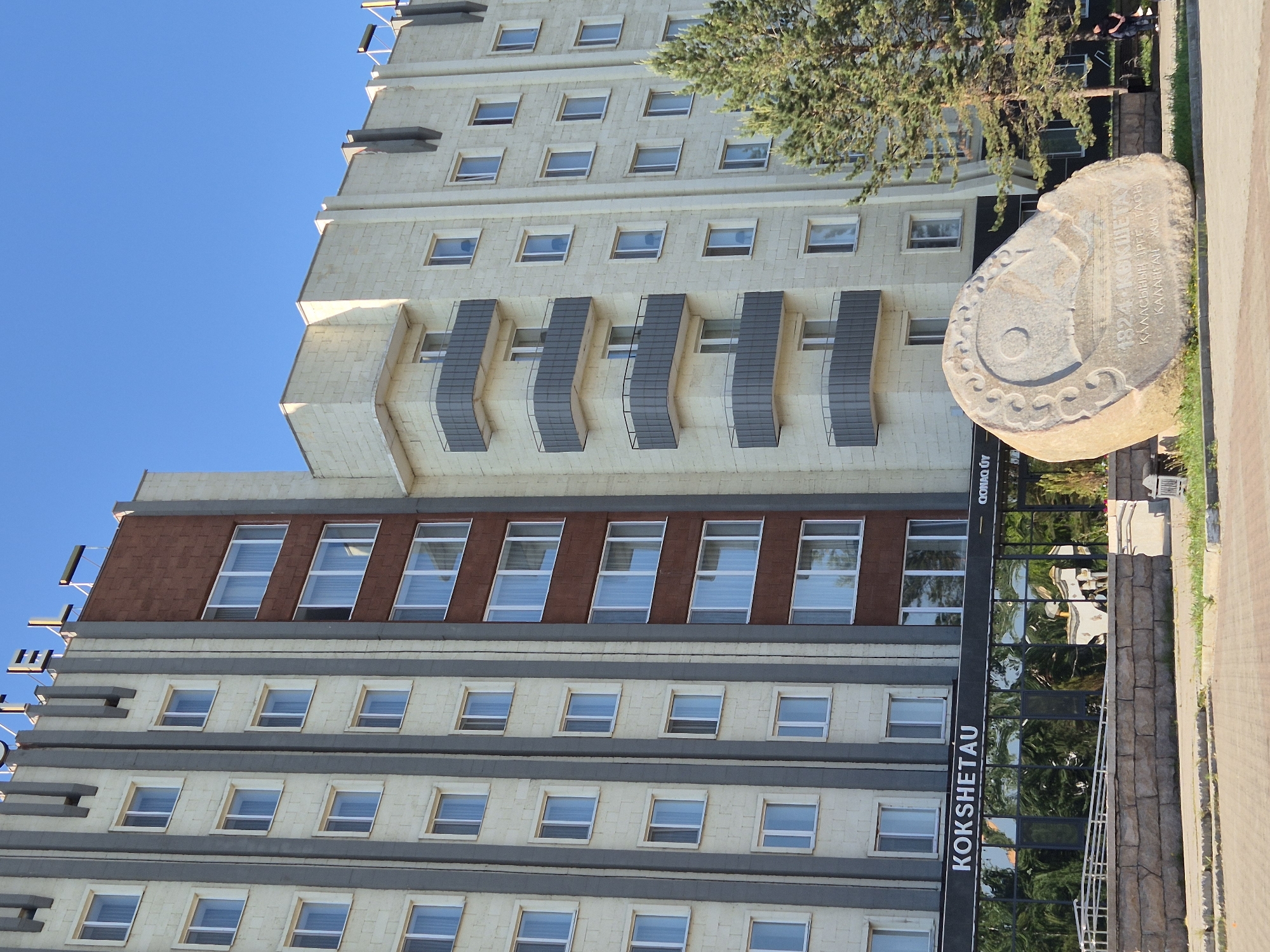 The image shows a multi-story building in Kokshetau, Kazakhstan. The building has a light-colored facade with rows of windows and some sections with reddish-brown brick. There are also some decorative elements such as protruding, curved balconies with railings. Adjacent to the building is a large, natural-looking stone with an inscription in Russian and Kazakh, commemorating the founding of Kokshetau in 1824. The entrance of the building has a glass facade with the name "KOKSHETAU" vertically written. A tree partially obscures the upper part of the building and stone. A paved walkway is visible next to the building, with a curb and green grass. A blue sky is visible above.