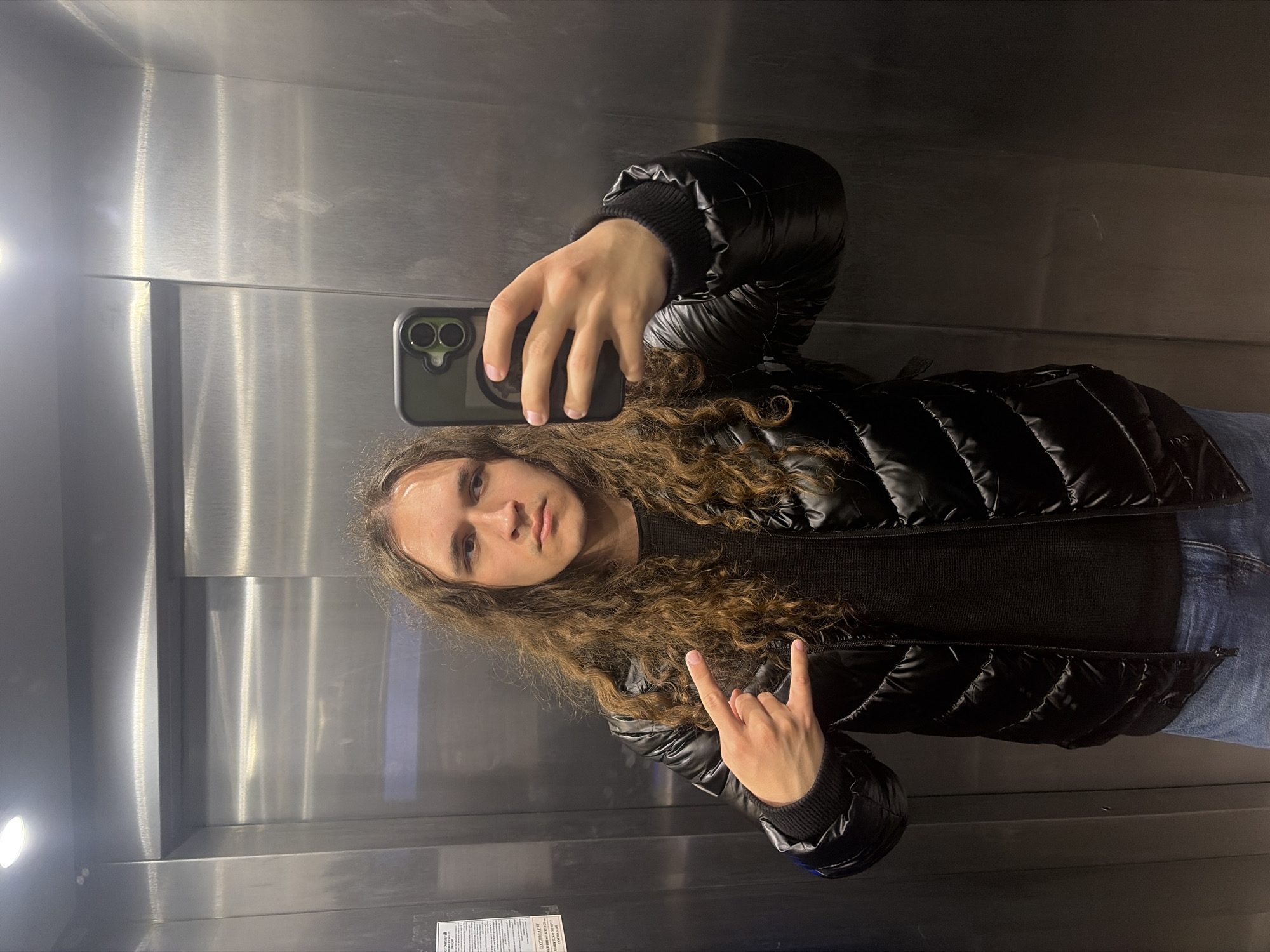 A young person with long, curly brown hair takes a mirror selfie inside an elevator, likely located in Plovdiv, Bulgaria. The individual, appearing to be male, is positioned centrally in the frame, holding a dark-cased iPhone with a dual-camera setup in their right hand to capture the reflection. They are wearing a black ribbed long-sleeved shirt under a shiny black puffer jacket, with blue jeans visible towards the lower right. With their left hand, they make a "rock on" gesture.
The elevator interior is characterized by brushed stainless steel walls, which reflect a bright light source on the left side of the frame. Vertical seams in the metal panels are noticeable in the background. The person's expression is neutral, bordering on serious. On the lower left wall, a small white rectangular sign with text, presumably in Cyrillic script given the location context, is visible. The scene captures a casual moment in a modern elevator setting.