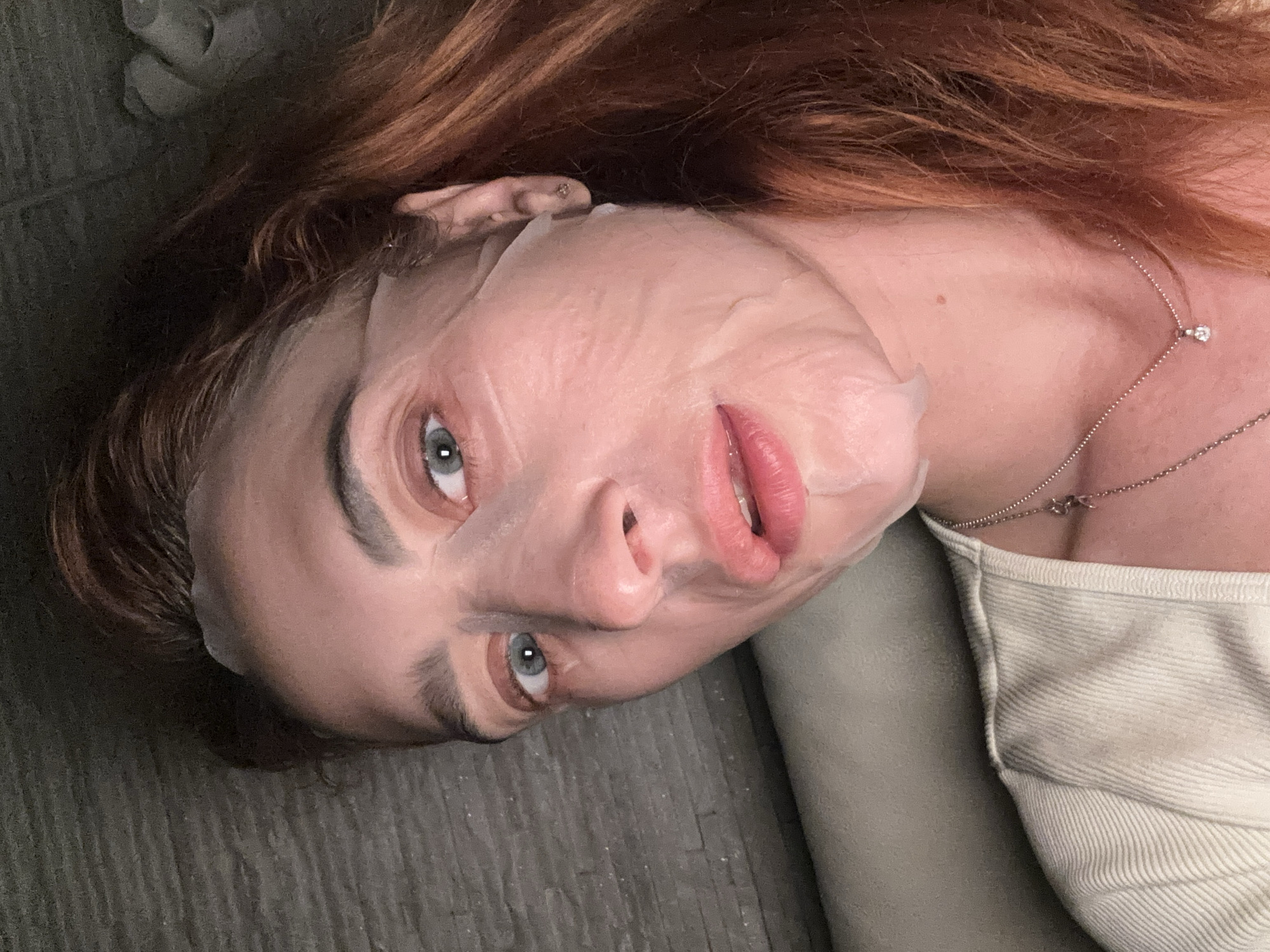 A woman lies on what appears to be a gray, slightly textured surface, possibly a floor or couch. She has reddish-brown hair and blue eyes. She is wearing a white shirt and a delicate silver necklace with a small, possibly diamond, pendant. She has a translucent face mask applied. The background is nondescript. The location of this photo is not discernible as Budva, Montenegro.