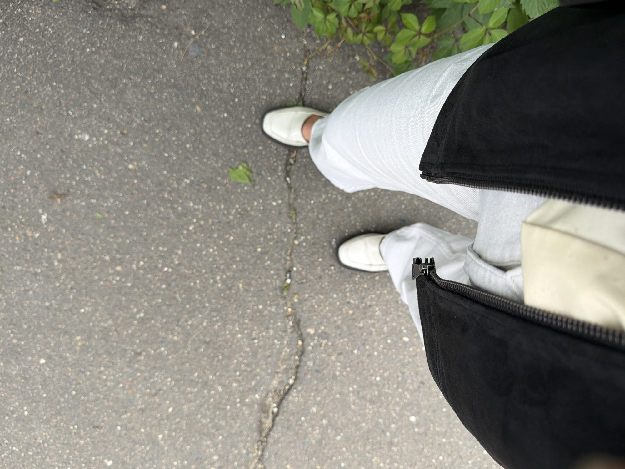 An overhead shot shows a person's legs and feet standing on a cracked asphalt surface. The person is wearing white loafers, light-colored pants, and a black jacket that is partially unzipped. The lower portion of the frame is filled with the texture of the asphalt, which appears somewhat rough and speckled. To the right of the frame, some green foliage is visible, suggesting an outdoor urban environment. The lighting is diffused, indicating an overcast day or a shaded area. The focus is on the clothing and footwear of the individual.