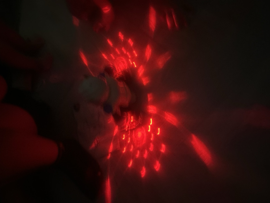 This image depicts a dimly lit indoor scene within the city of Chaikovsky, Russia. At the approximate center of the frame is an indistinct, light-colored object or figure, possibly a small toy, exhibiting a faint blue detail on its upper portion. This central object is surrounded by, and appears to emit, a dense radial pattern of numerous small, intensely bright red lights. These lights manifest as distinct, short, often crescent-shaped or rectangular segments, generating prominent red light streaks and a pervasive red glow that illuminates the immediate environment. The background is predominantly obscured by darkness, with some blurry red forms visible in the upper portion of the frame, suggestive of reflections or other out-of-focus objects. No individuals or specific actions are discernible within the scene.