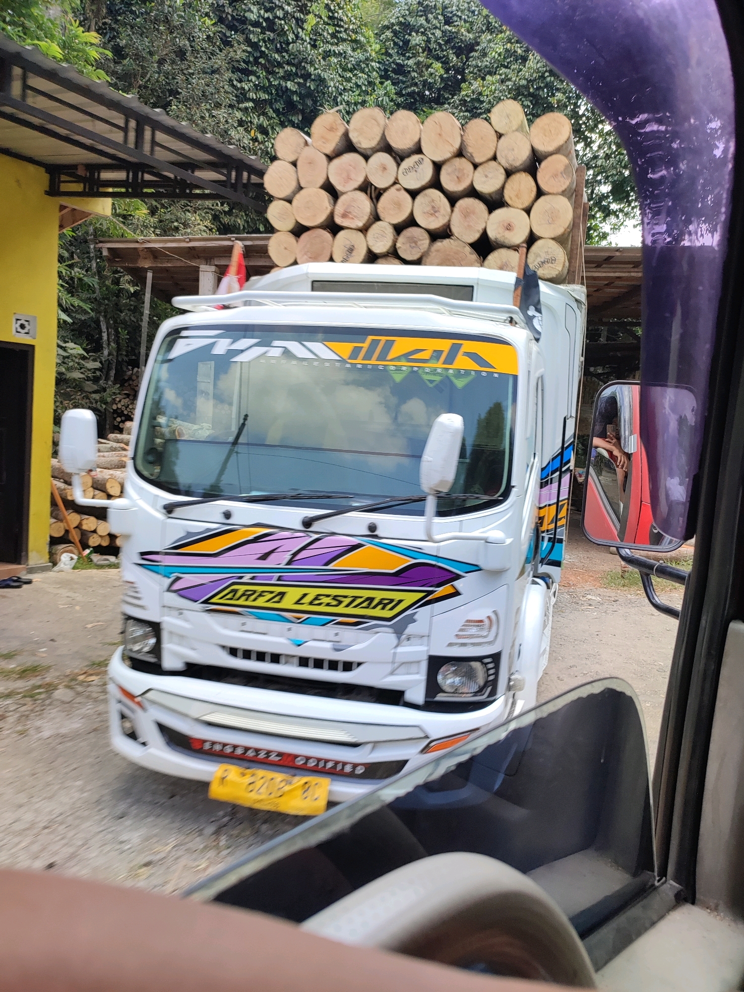 A white truck loaded with logs is parked on a dirt road in front of a yellow building with a metal awning. The truck has colorful graphics on its side and front, and a license plate with the characters "P 8208 BC". A person is visible in the side mirror of another vehicle, and trees and foliage are in the background. The scene appears to be in a rural or semi-rural area.