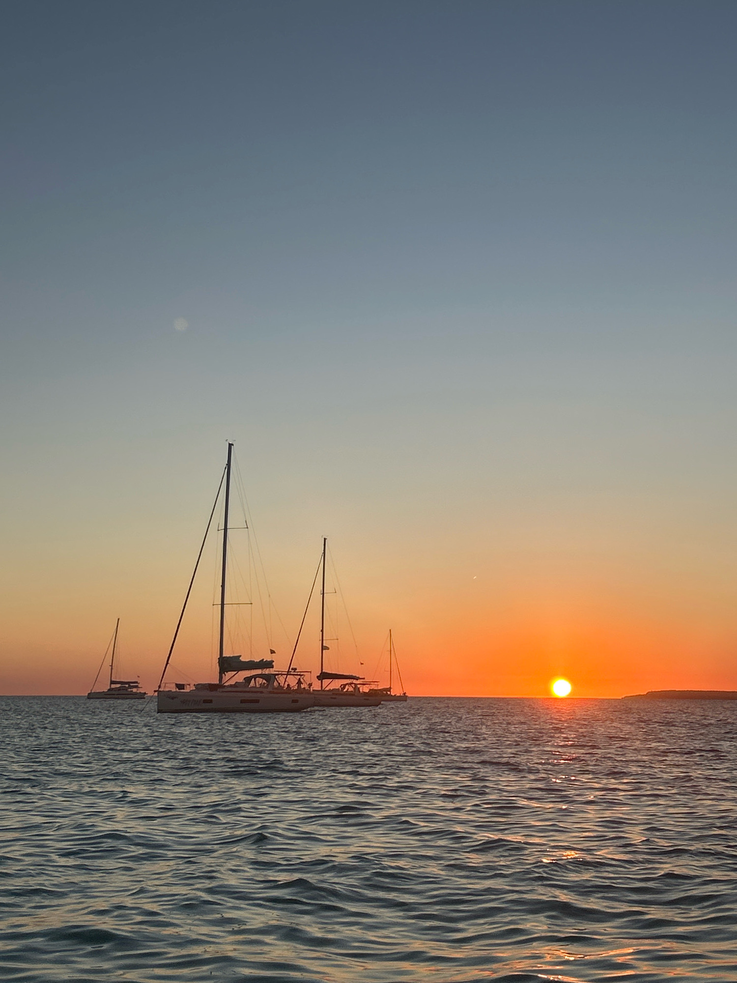 The image depicts a sunset over the sea in Campos, Spain. The sky transitions from a pale blue at the top to a vibrant orange near the horizon. A bright, round sun is visible just above the sea. Several sailboats are present on the water. In the foreground, the water is rippled, reflecting the colors of the sunset. The overall scene is serene and picturesque, characteristic of coastal sunsets.