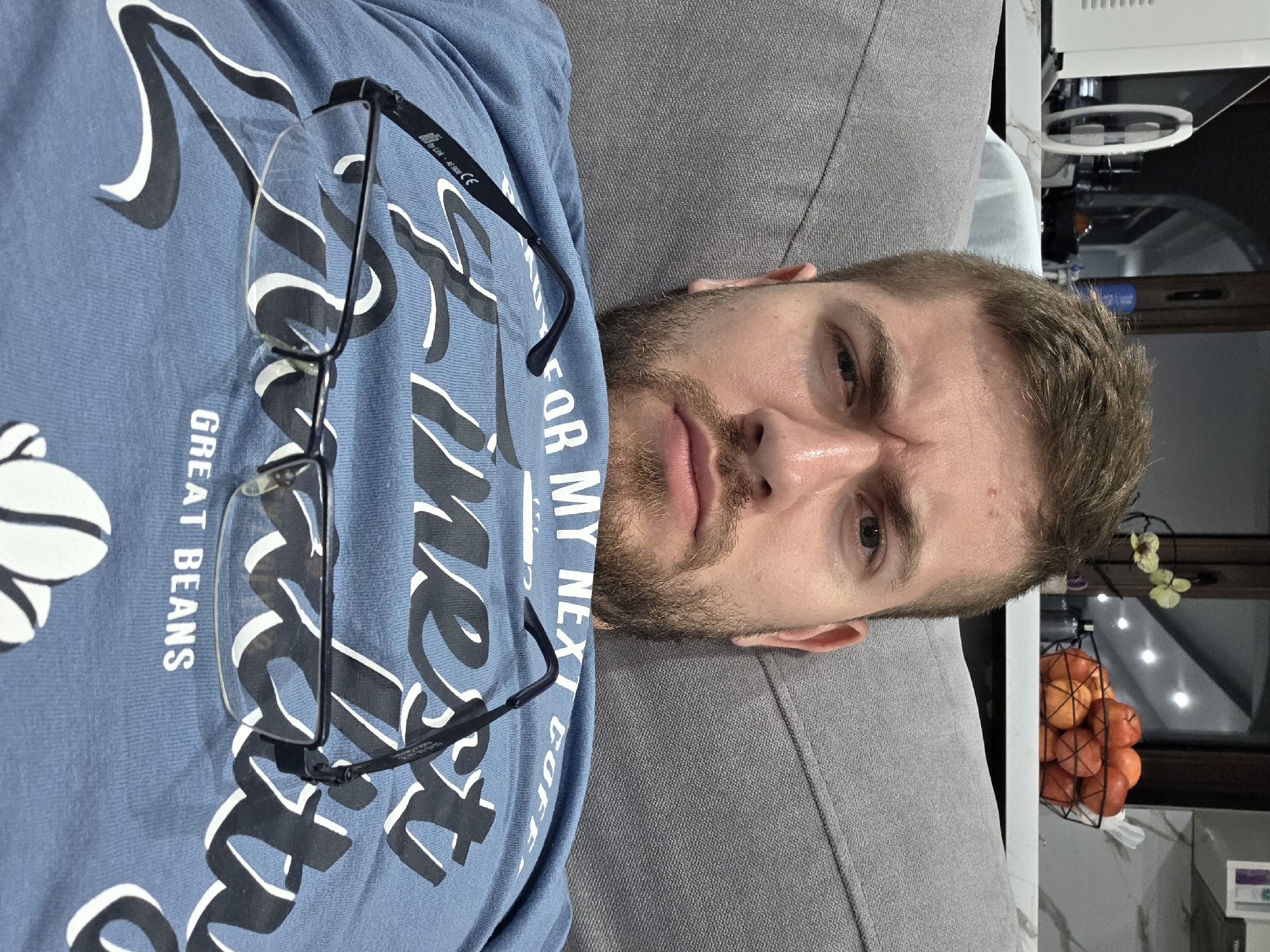 A man with short blonde hair and a beard lies on a gray couch wearing a blue t-shirt with white graphics and the words "GREAT BEANS" and "FOR MY NEXT COFFEE." A pair of black-rimmed glasses rests on his chest. Behind him is a white kitchen appliance and a wire basket filled with oranges. There is no verifiable evidence to suggest that this image was taken in the city of Yagodovo, Bulgaria.
