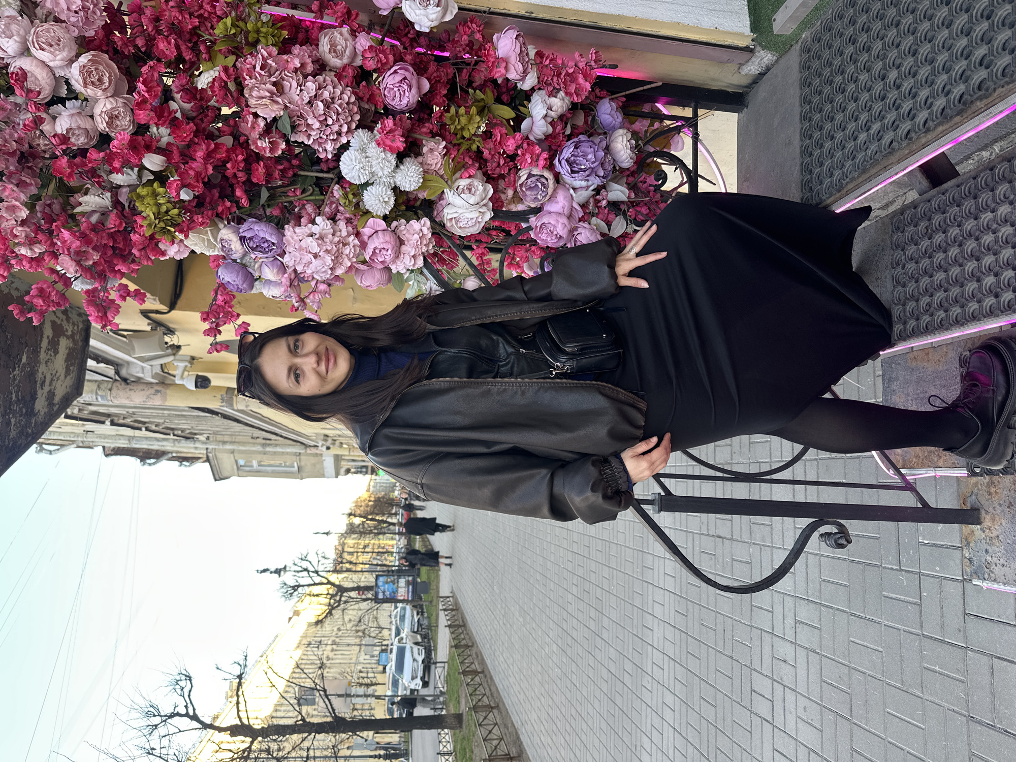 A woman is seated on a black metal decorative railing, positioned in front of a vibrant display of artificial flowers. The flower arrangement features various shades of pink, magenta, purple, and white blooms with some green foliage, possibly adorning the exterior of a building or a large planter.
The woman has long dark hair, parted in the middle, with sunglasses perched on top of her head. She is wearing a dark leather jacket, a dark top (possibly blue underneath), a long black skirt, dark tights or leggings, and dark, robust boots. Her left hand rests on her lap, while her right hand is near her waist. She looks directly at the camera with a calm, subtle smile.
Behind her, the scene extends to a paved sidewalk, indicating an urban setting. Bare trees line the street, suggesting cooler weather or a late autumn/winter season. Buildings with classic architectural styles are visible in the background, and several cars are parked along the street. A few indistinct figures can be seen walking in the distance. The sky above appears bright, suggesting daytime, though the light is soft rather than harsh sunlight. Security cameras are visible near the flower display. There is no discernible text visible in the image. The setting appears to be a street in Saint Petersburg, Russia.