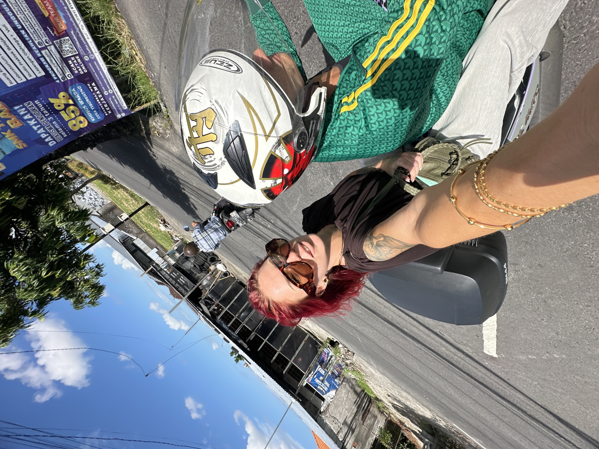 Here is a detailed description of the image:
The image is a selfie taken on a road in North Kuta, Indonesia. The person taking the selfie has red hair, sunglasses, and gold bracelets. A motorcycle helmet is visible on the person riding the motorcycle, suggesting they are a passenger. The driver of the motorcycle is wearing a green Adidas jacket.
In the background, there are buildings and trees, with a blue sky and some clouds visible above. There is a sign on the left side of the image that appears to advertise a school called SMK TI Bali Global Badung. The sign mentions educational programs and offers 85% off for residents of Desa Tibubeneng.