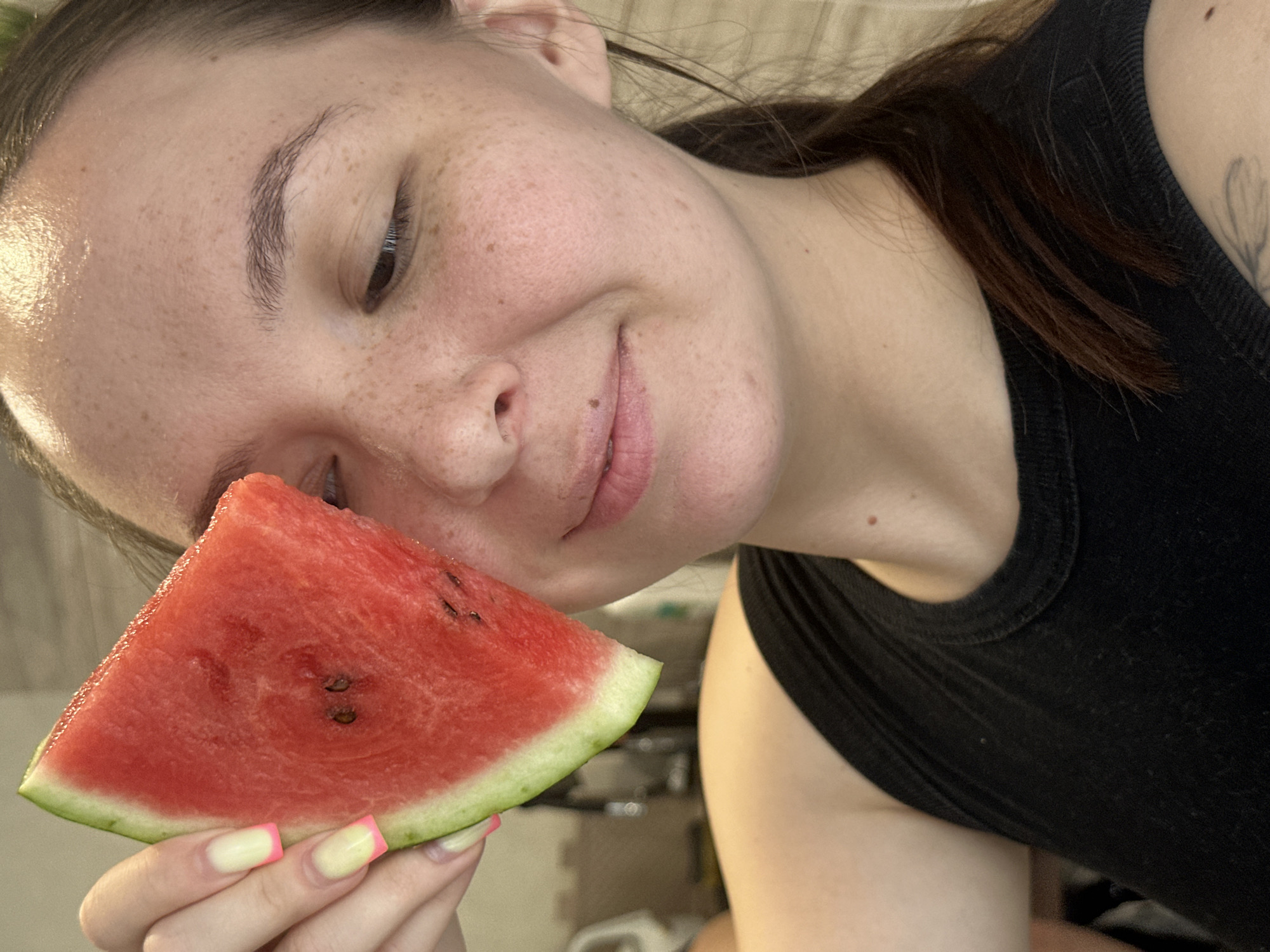 A close-up shot shows a young woman from the chest up, holding a slice of watermelon to her face. Her head is tilted downwards and to the left, with her eyes looking down and a slight smile on her lips. She has freckles across her nose and cheeks. Her nails are painted a pale yellow with pink tips. She is wearing a black tank top. The background is blurred, but appears to be indoors with neutral-colored walls and a hint of something light-colored in the lower right corner. The lighting suggests daytime.