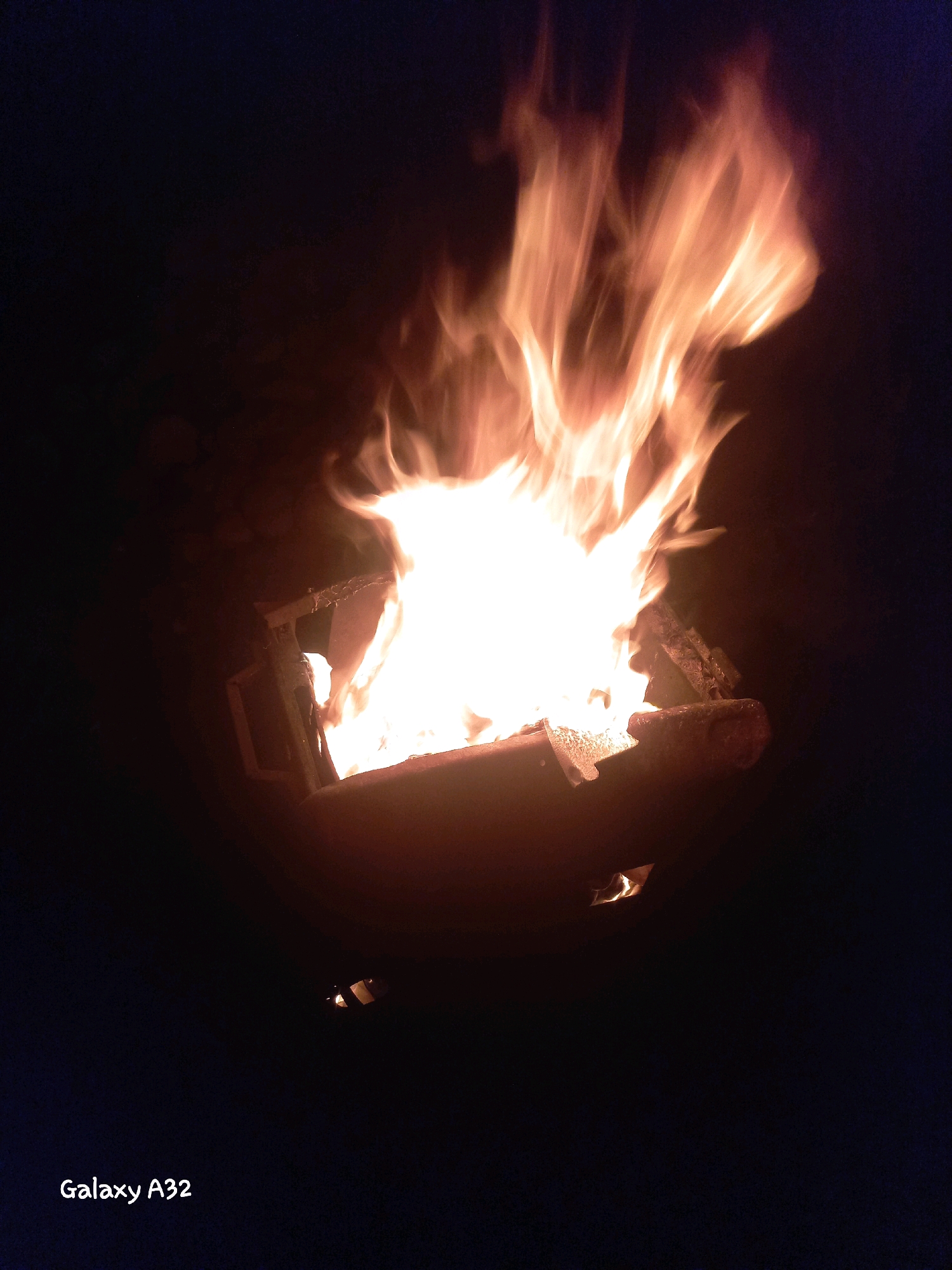 A fire burns in a dark, outdoor setting. The flames are bright orange and yellow, reaching upwards from a circular metal container that appears to be a fire pit. The background is dark, suggesting it is nighttime. The lower left corner of the image contains the text "Galaxy A32," indicating the device used to capture the photograph. The location is Kemin, Kyrgyzstan.