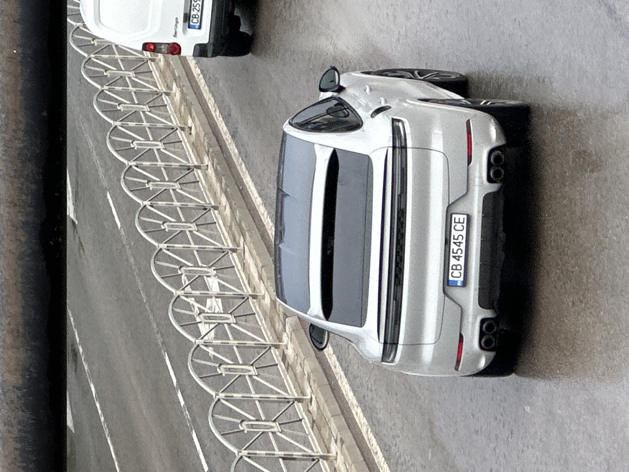 The image, rotated 90 degrees clockwise, captures a daytime street scene in what appears to be an urban or suburban environment. The main subject is a silver (or light grey) Porsche Cayenne Coupe, parked facing away from the viewer on a paved surface. The car exhibits a sleek design with a sloping roofline, black-tinted rear windows, and two prominent circular exhaust tips on either side of the rear bumper. Its Bulgarian license plate displays "CB 4545 CE".
To the left of the Porsche, a distinctive light-colored metal barrier, featuring a series of semi-circular sections with spoked patterns, runs parallel to the road. Beyond this barrier, a paved road with white lane markings is visible. A white Citroën Berlingo van is partially visible further down the road, behind the barrier. Its rear section shows the word "Berlingo" and part of a Bulgarian license plate, starting with "CB 259".
On the far left, a dark, thick vertical object, possibly a pole or pipe, frames the edge of the scene. The weather appears clear with good natural lighting. No people are present, and both vehicles are stationary. The Bulgarian license plates confirm the location context of Veliko Tarnovo, Bulgaria.