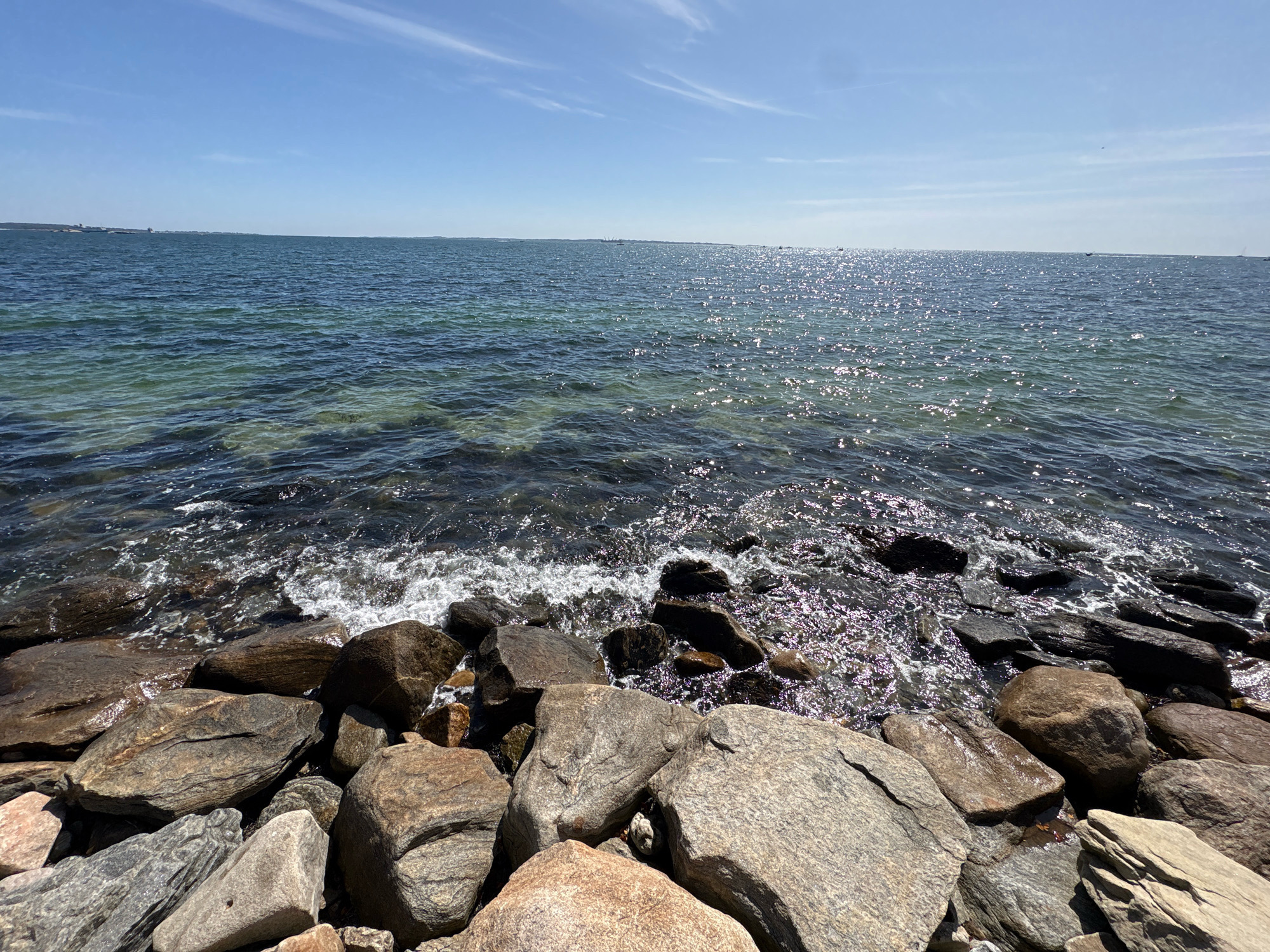 A wide shot captures a rocky shoreline meeting the ocean under a clear blue sky. The ocean is a deep blue with patches of lighter turquoise near the shore, indicating shallow water. Sunlight glints off the water's surface, creating a shimmering effect. Small waves crash against the dark, wet rocks and pebbles that form the foreground. Beyond the water, a distant, hazy landmass is visible on the horizon, dotted with structures. The sky is a brilliant blue, with a few wispy white clouds. The image appears to be taken from a vantage point slightly above the water level, looking out towards the open sea. The location is identified as Waterford, United States of America, suggesting a coastal setting within that city. No people or animals are present in the frame.