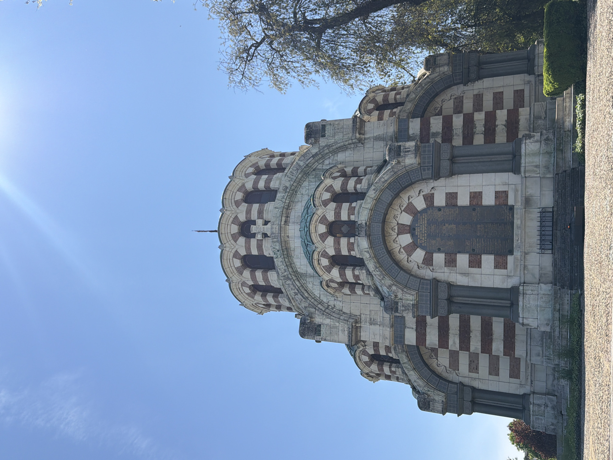 The image shows the Pleven Panorama, a monument in Pleven, Bulgaria, built in 1977 to commemorate the Siege of Pleven in 1877. The monument is a cylindrical structure with alternating white and reddish-brown stripes. It is comprised of multiple levels with arched openings. The bottom level has a dark plaque and pillars. A tree and a well-manicured hedge are visible to the right of the monument. The sky is clear blue with a bright light source in the top left corner.