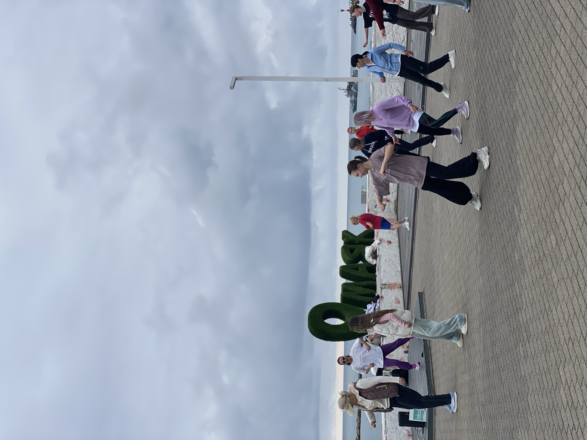 A group of people, mostly young women, are participating in an exercise or dance class on a paved promenade. The class is taking place next to large, green, topiary letters that spell out "СОЧИ", which indicates the city of Sochi, Russia. The participants are dressed in casual athletic wear, and are arranged in a line along the promenade. A tall white lamppost stands nearby. In the background, the sky is overcast and a body of water can be seen, likely the Black Sea.