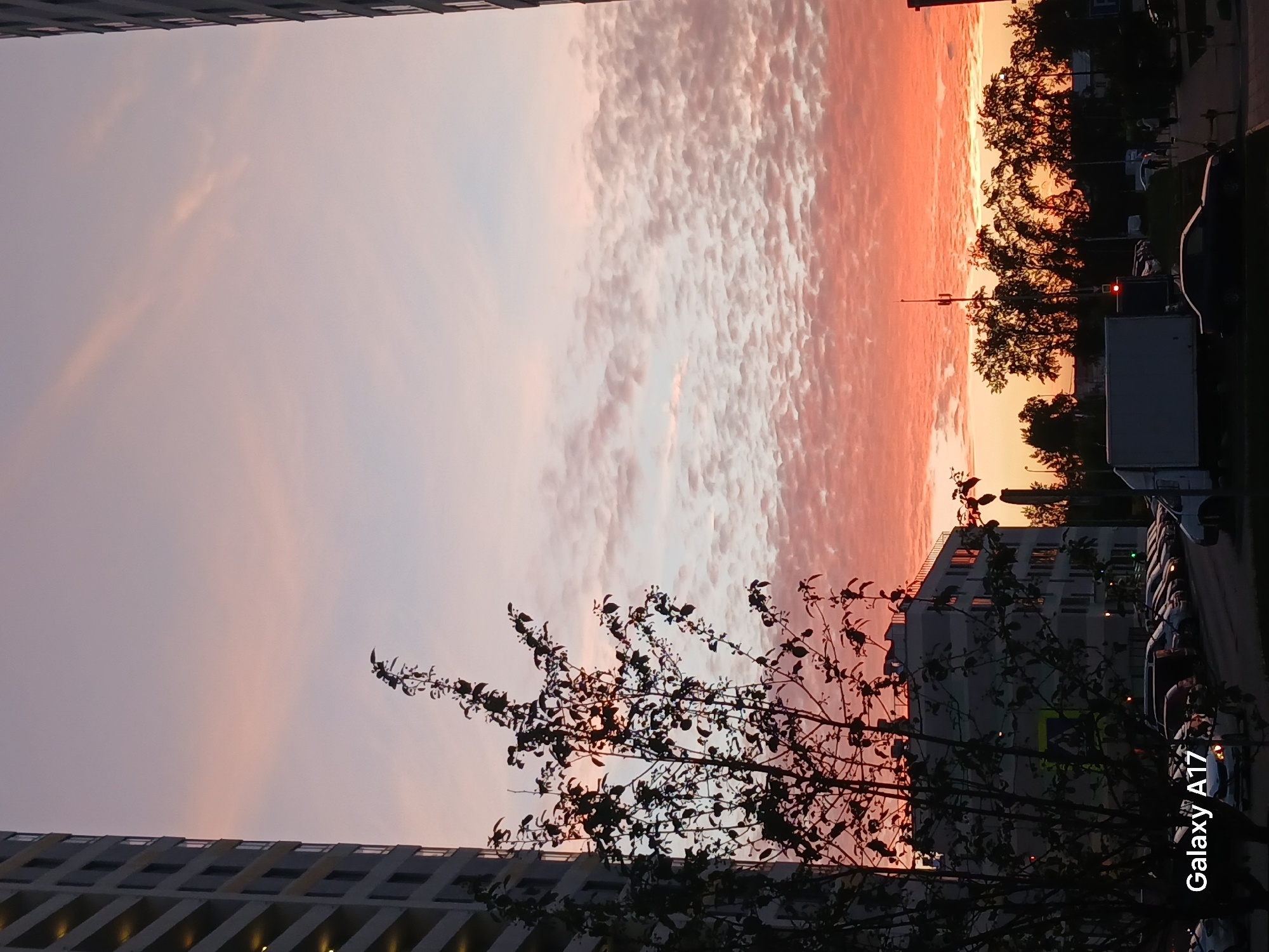 The image captures a sunset scene in a city, likely Yekaterinburg, Russia, based on context. The sky exhibits a gradient from pale purple to a vivid orange-red hue near the horizon, with textured clouds spanning the sky. The foreground contains a silhouetted tree with leaves, and buildings on either side of the frame, indicating an urban setting. The right side of the image reveals a street with parked cars and additional buildings in the background, including what appears to be a delivery truck. A mobile phone watermark "Galaxy A17" is present in the bottom right corner of the image. No people are visible.