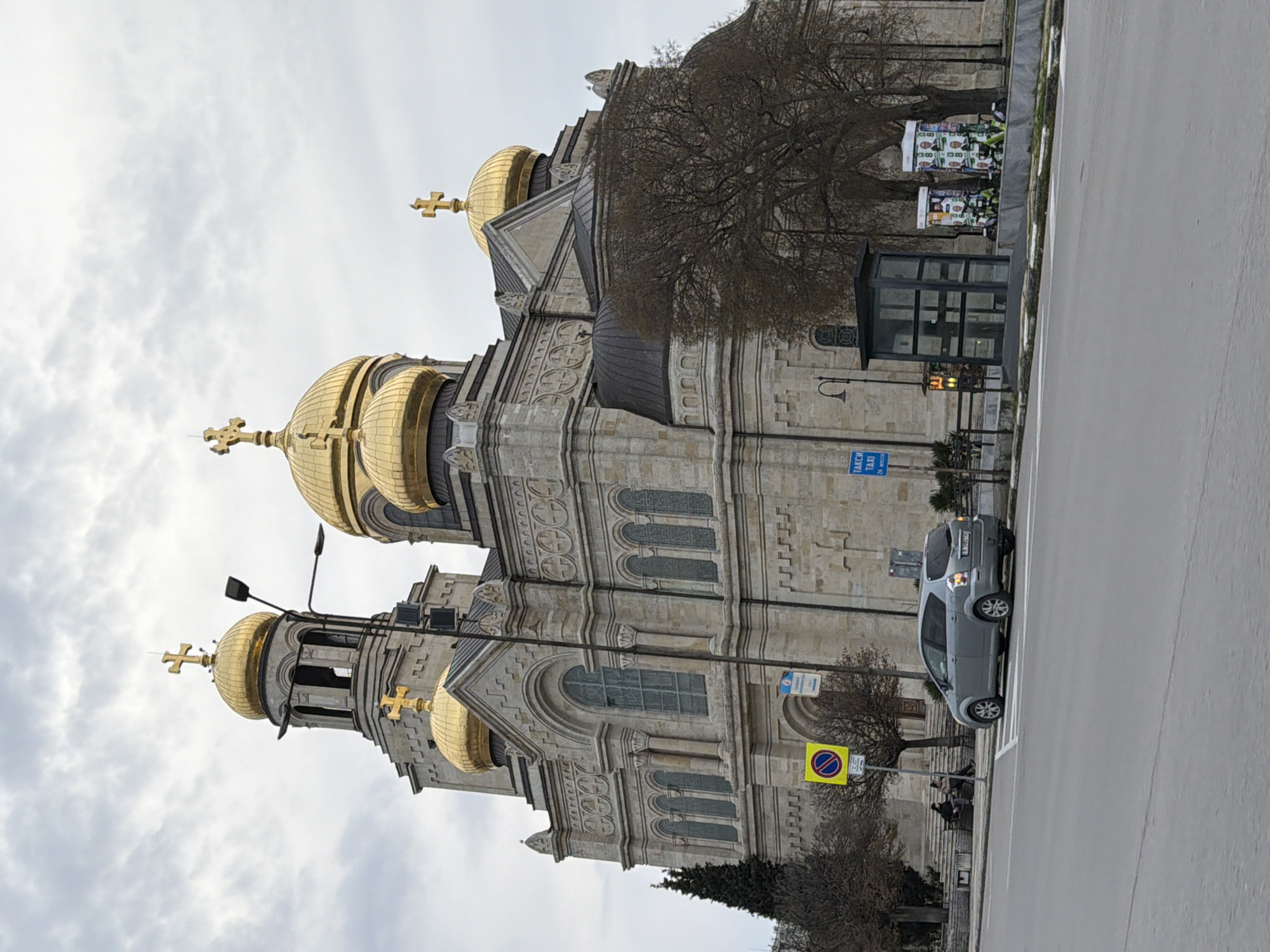 The image shows the Cathedral of the Assumption of the Virgin in Varna, Bulgaria. The cathedral is a large stone building with golden domes and crosses. A silver car is parked on the street next to the cathedral. A bus stop and a no-parking sign are visible. Several people are sitting on the stairs. The sky is cloudy.