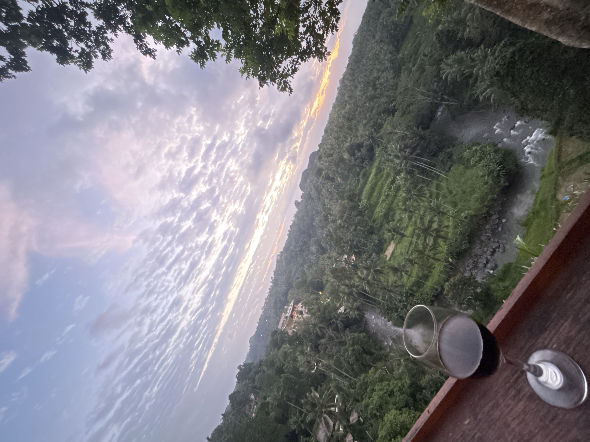 A glass of red wine sits on a wooden railing overlooking a lush, tropical valley. The valley is filled with dense green vegetation, including palm trees and rice terraces, and a winding river flows through it. In the distance, a few buildings are visible, nestled among the trees. The sky above is a soft pastel palette of pink, purple, and blue, with wispy clouds creating a textured canvas. The sun appears to be setting or rising, casting a warm glow on the horizon. The scene suggests a serene and picturesque setting, likely in a place like Ubud, Indonesia, known for its natural beauty and tranquility.