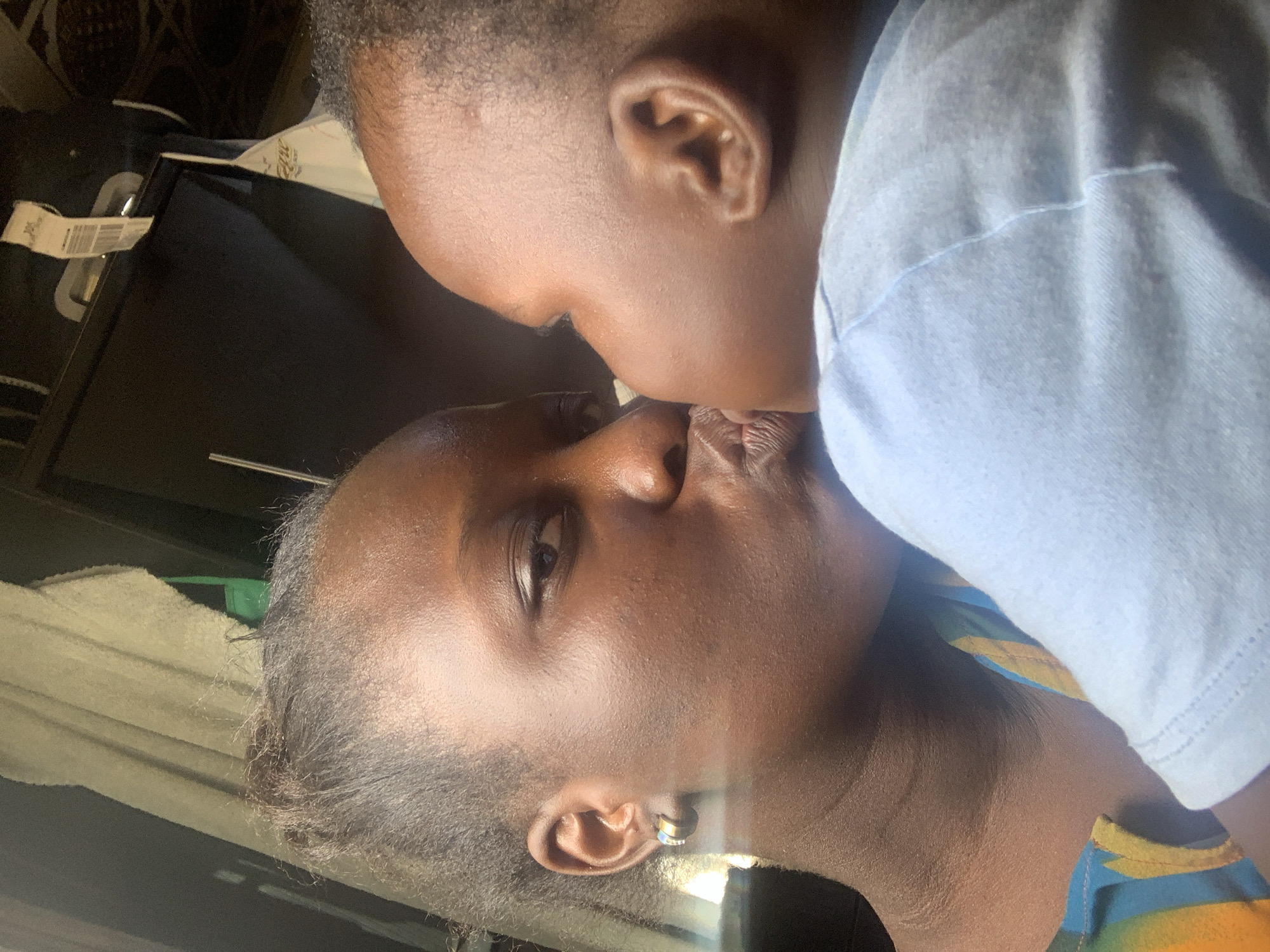 A close-up, overhead shot shows two individuals, presumably a mother and child, sharing an intimate moment. The mother, on the left, is in profile, her eyes closed as she leans in to kiss the baby on the right. The baby's face is also in profile, with their eyes open. Both have dark skin and are wearing casual clothing. The background is out of focus, but appears to be an indoor setting with soft lighting. The overall impression is one of warmth and affection.