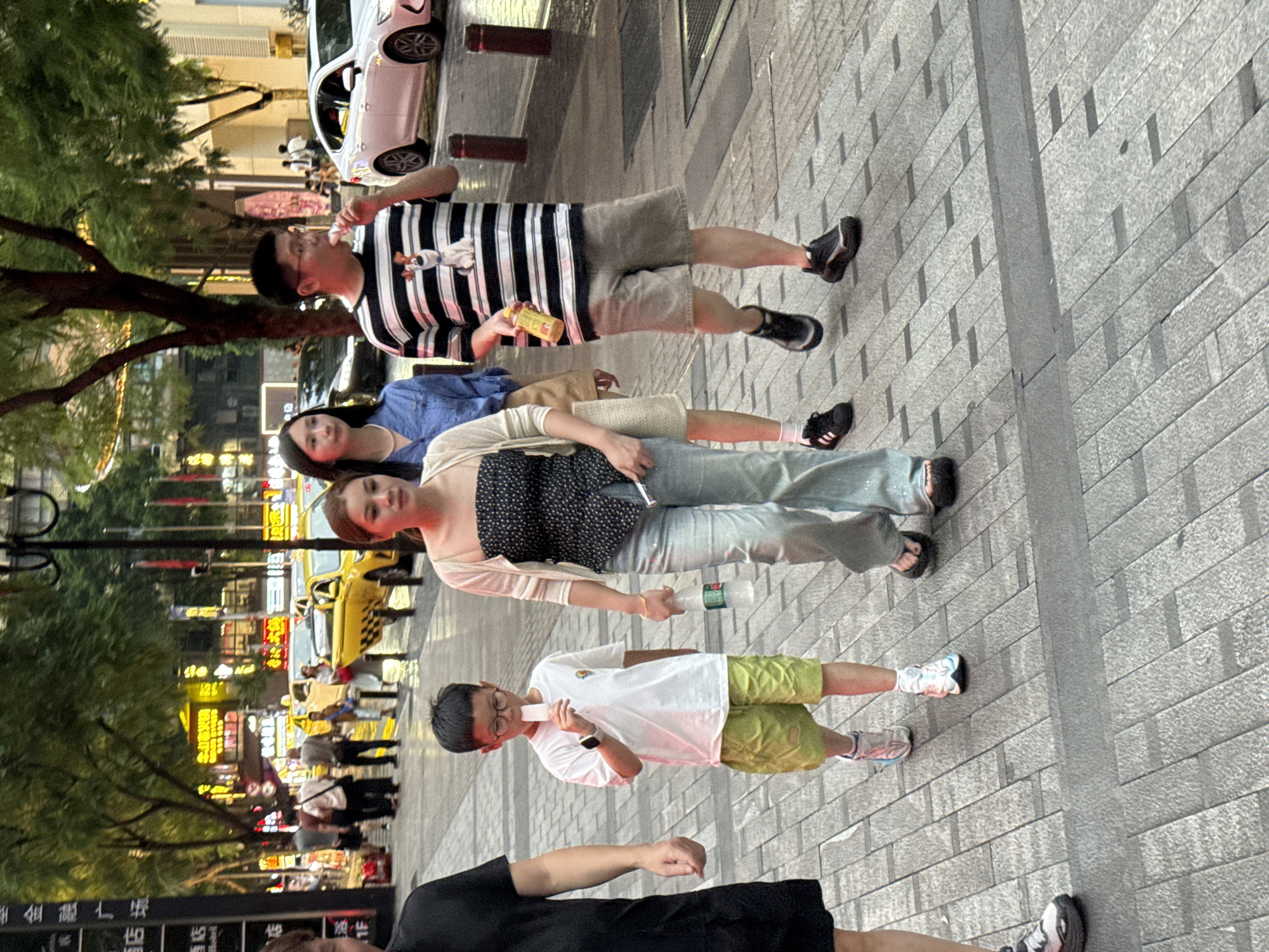 In the Yuzhong District of China, on a city sidewalk, a group of people are walking. A young boy in a white t-shirt and green shorts is eating an ice pop. To his right, a woman in a black and white patterned top and a light-colored cardigan is holding a water bottle. Behind her, a woman in a blue denim shirt walks with a light brown purse. To the right of the boy, a man in a black and white striped t-shirt and grey shorts is eating an ice pop and holding a yellow bottle. The sidewalk is paved with grey rectangular tiles. In the background, yellow taxis are visible on the street, along with trees and illuminated buildings. The overall scene depicts a casual urban streetscape.