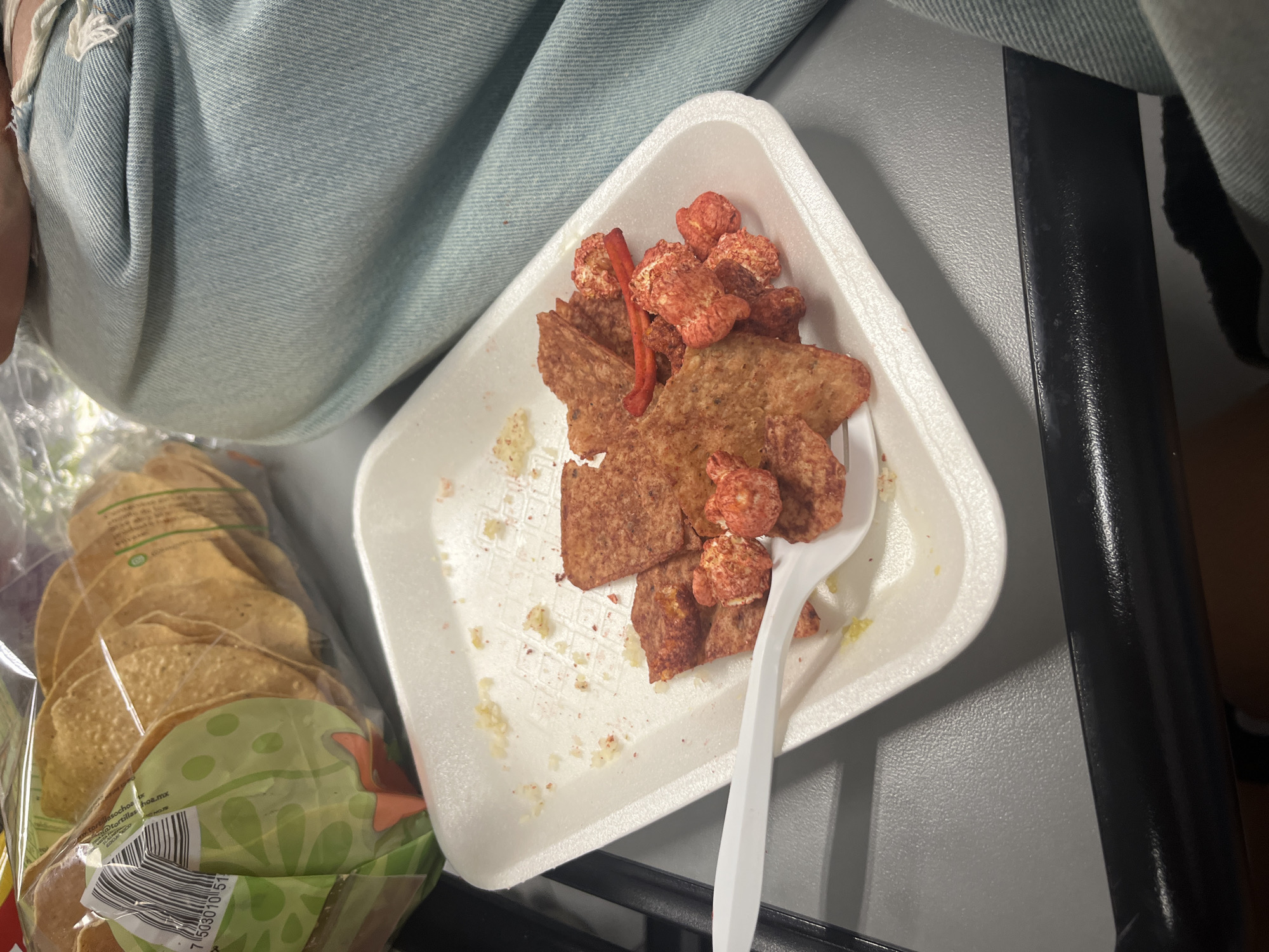 The image shows a snack tray on a gray surface, possibly a table or tray table. The tray is white and triangular, containing what appears to be tostada chips and puffed corn snacks, seasoned with a reddish powder. A single red, thin, curly piece of chili pepper is present. There is a white plastic fork on the tray. A clear plastic bag of tortillas is also visible next to the tray. The background includes the lower part of a person's leg, clad in light blue denim jeans.