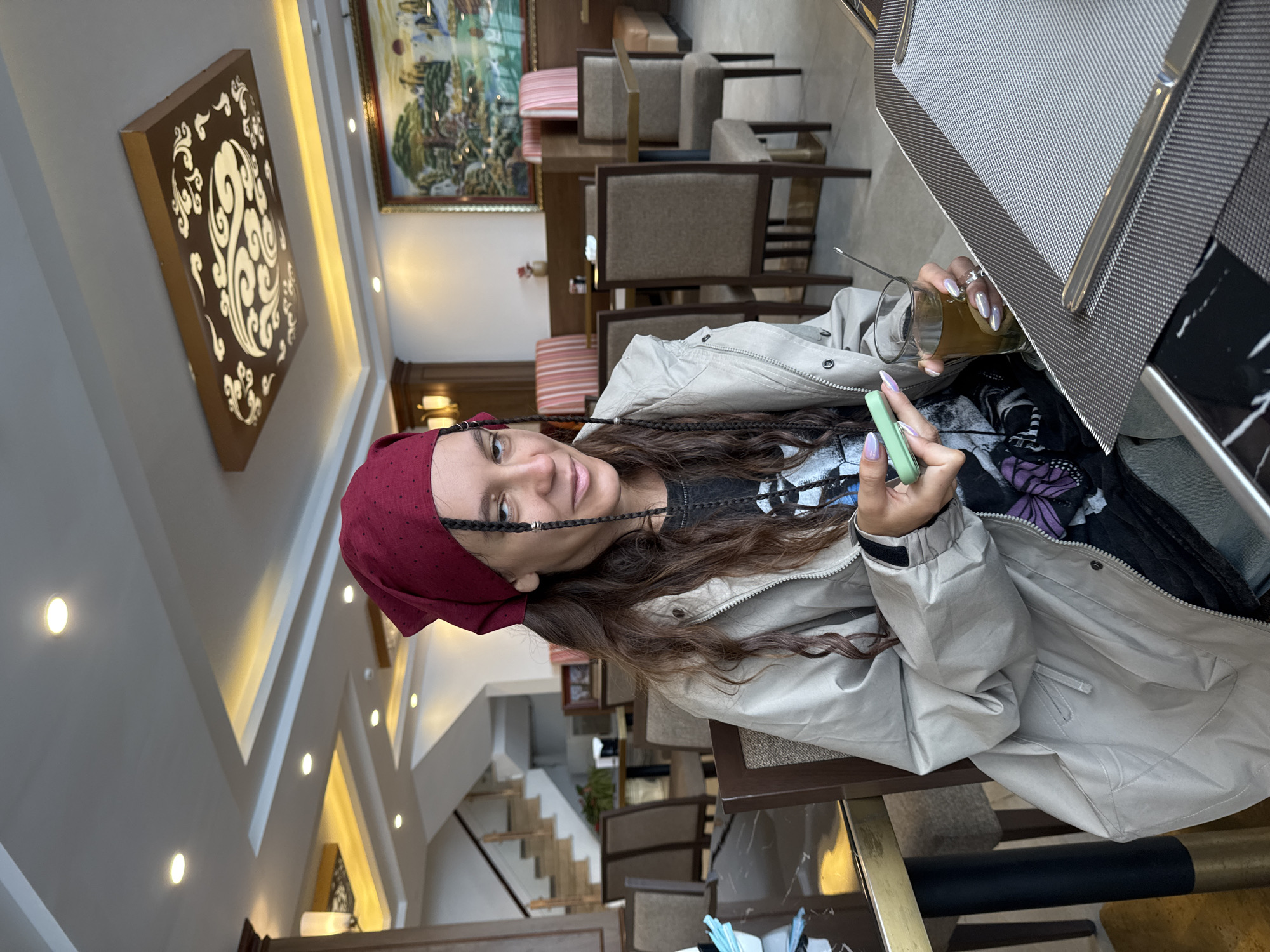 The image shows a woman in what appears to be a restaurant. She is wearing a maroon headscarf and a light beige jacket. She has dark hair with braids. She is holding a green phone in one hand and a glass with liquid in the other. She is sitting at a table. The restaurant has neutral-toned chairs and tables, with a decorative painting hanging on the wall. There is also a decorative panel on the ceiling. There are stairs in the background. The image does not confirm a specific location in Binh Lu, Vietnam, but the setting suggests an indoor dining area in that city.