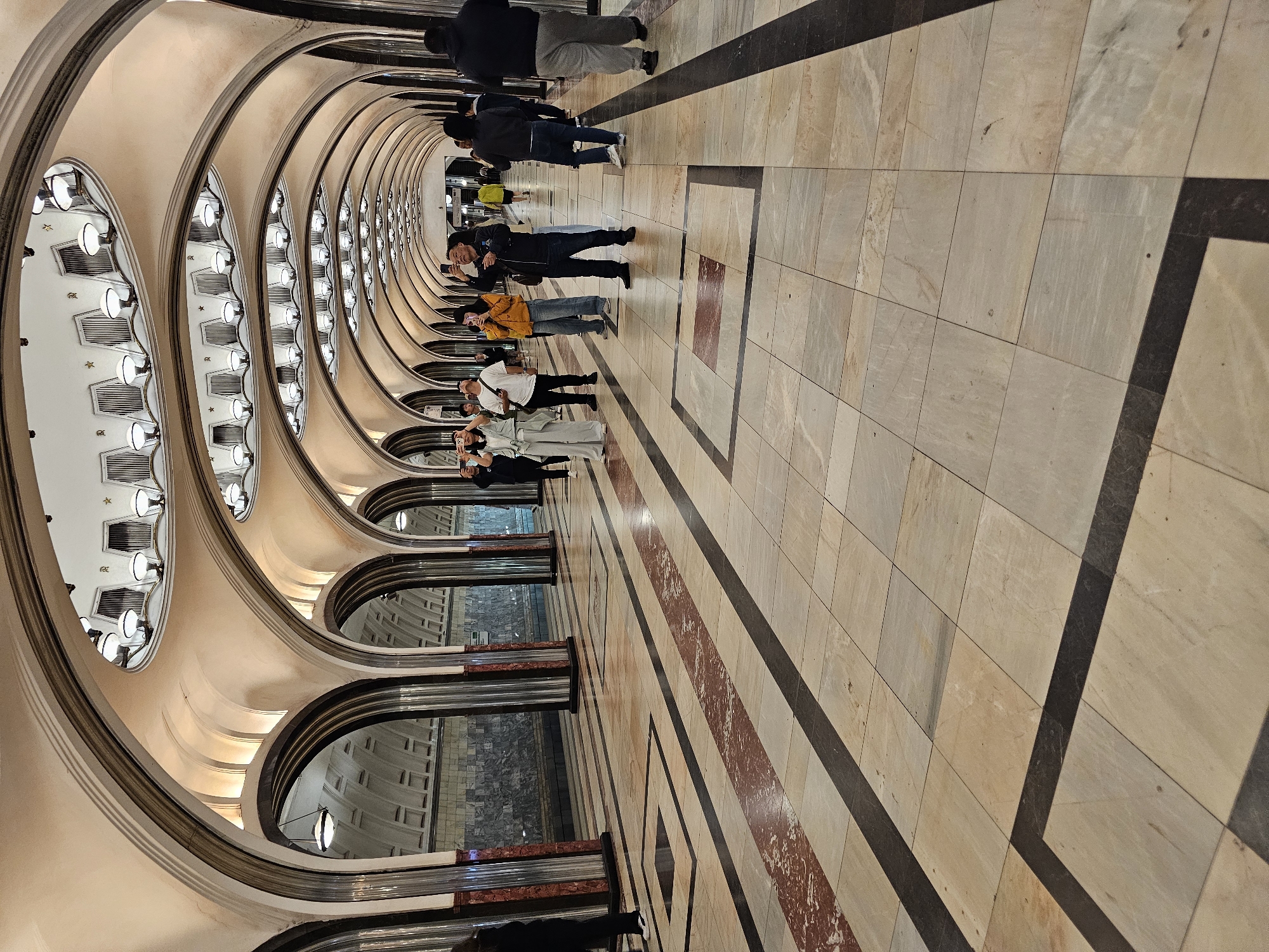 The image depicts the vestibule of a Moscow Metro station, likely Mayakovskaya Station, given its distinctive architecture. The station features a long, arched corridor with a high, vaulted ceiling adorned with rows of bright, circular lights. The walls are lined with marble and ornate mosaics, and the floor is paved with large, polished stone tiles in a geometric pattern of beige, white, and dark brown with red accents. Several people are visible walking down the corridor, many of whom are looking up and taking photographs with their smartphones, suggesting they are tourists or admiring the station's beauty. The perspective emphasizes the grandeur and symmetry of the architecture.