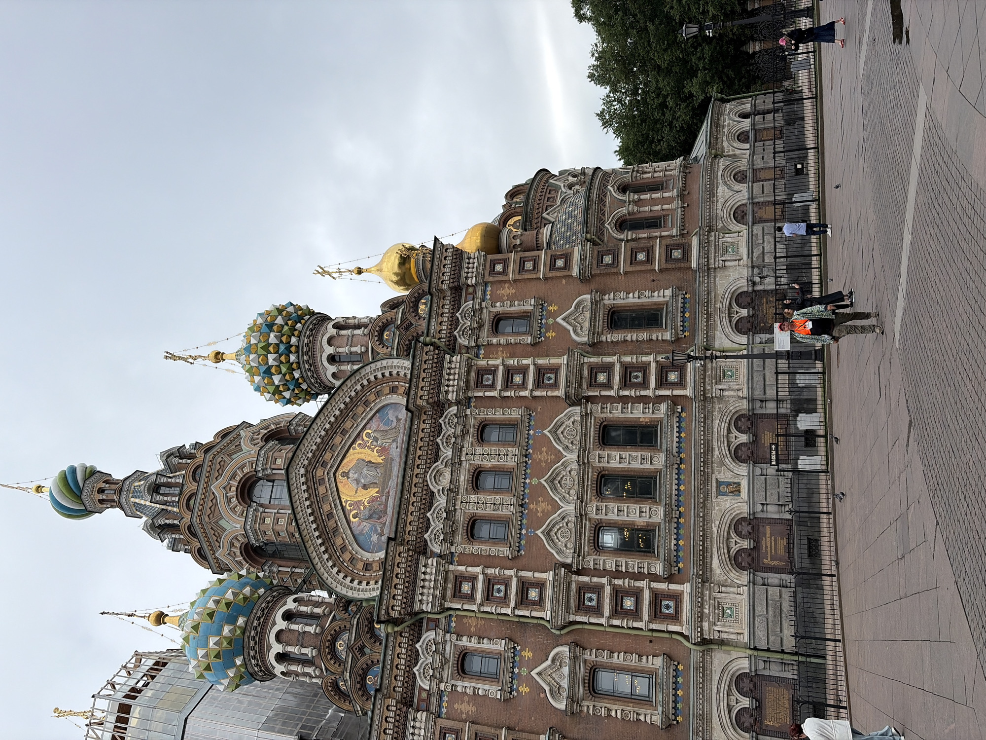 The image shows the Church of the Savior on Spilled Blood in Saint Petersburg, Russia. The ornate cathedral is captured from a low angle, emphasizing its elaborate architecture with multiple domes, intricate mosaics, and detailed facades. Several people are visible in the foreground and midground, some walking on the paved square in front of the church, while others are near the fence that encloses part of the building. The sky is overcast, suggesting a cloudy day, possibly in the morning or afternoon given the ambient light. The scene appears to be a typical day with visitors and locals moving around the iconic landmark. There is no visible text in the image.