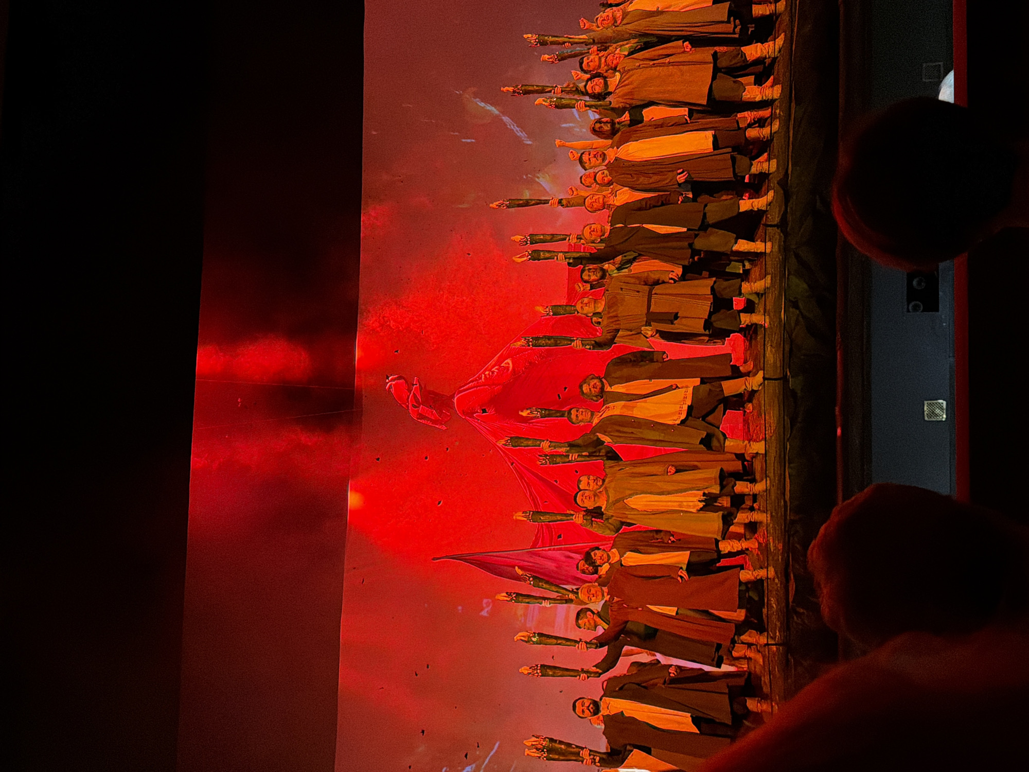 A group of people on a stage are holding up torches. The stage is lit with red lighting and a projection of a figure on a wire is visible in the background. The people are dressed in long robes and some have beards. They appear to be actors or performers in a play or musical. The image was taken from the audience, with the backs of two audience members visible in the foreground. The location is not specified in the image.