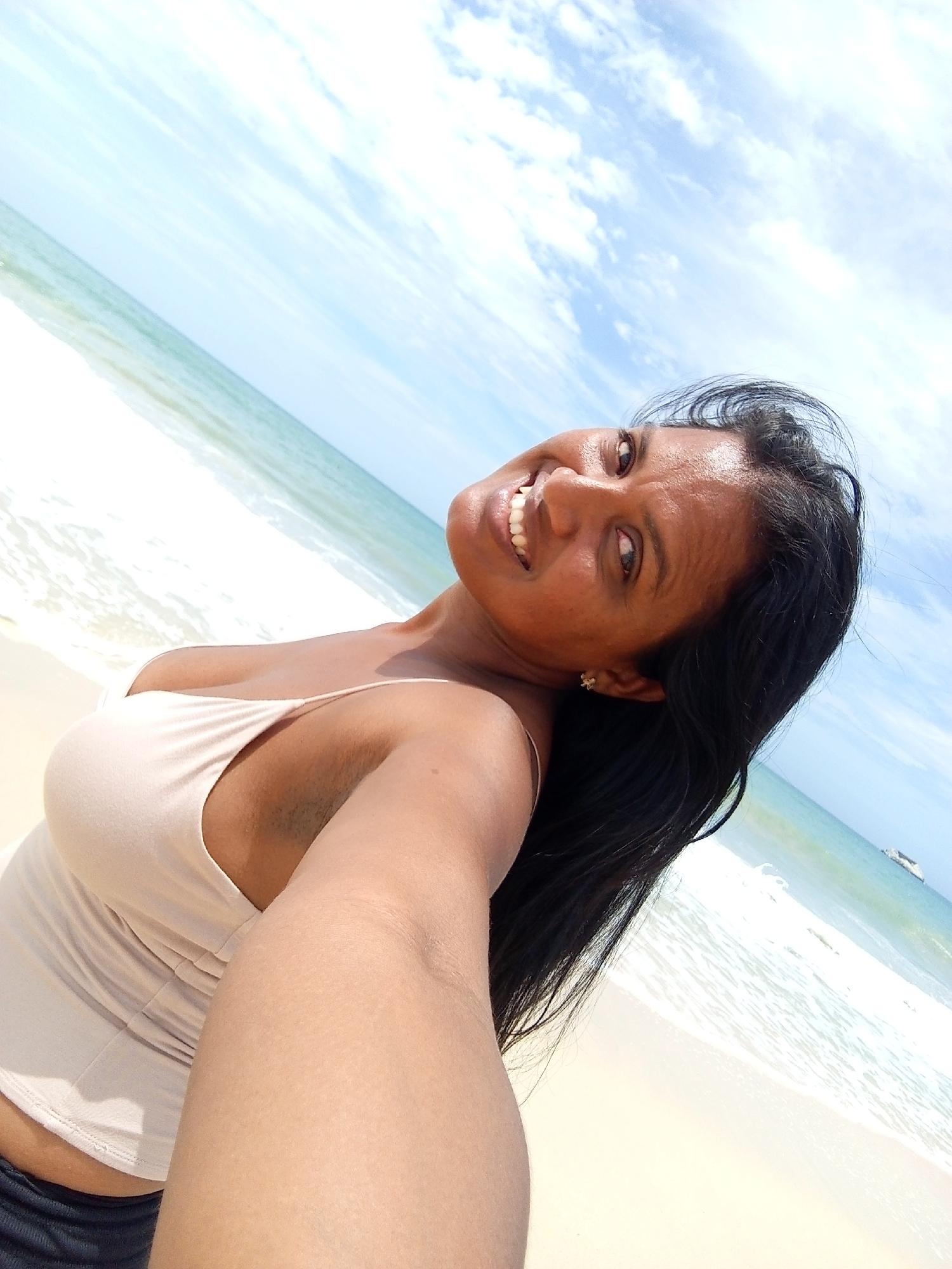 A woman is taking a selfie on a beach. The background includes a sunny sky with some clouds, the ocean with waves, and the sandy beach. The exact location in Altagracia, Venezuela, cannot be determined from this image.