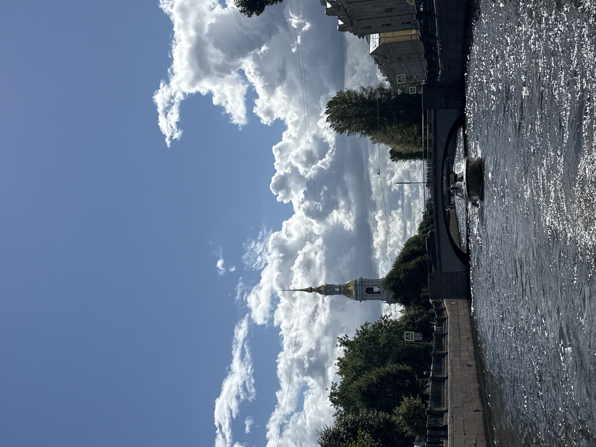This image depicts a sunny day with dramatic clouds over a canal in Saint Petersburg, Russia. In the background, a tall, slender bell tower with a golden spire rises above lush green trees. A stone bridge arches over the canal, and a white motorboat carrying two people navigates the dark, rippling water. The canal's stone embankments are visible on both sides, lined with trees and buildings. The overall scene conveys a picturesque urban landscape with a mix of historical architecture and natural elements.