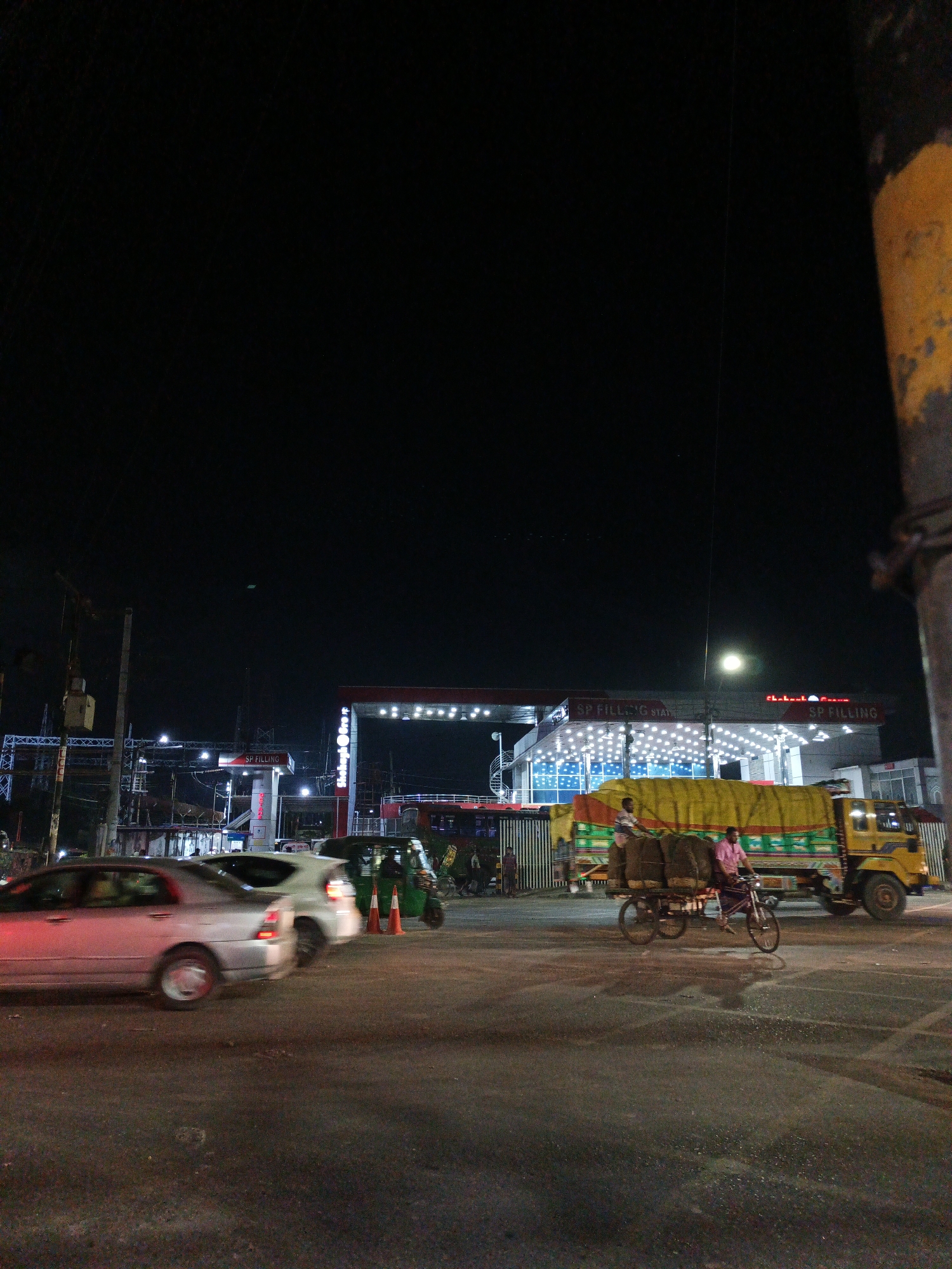 At night in an urban area of Unknown, Unknown, a brightly illuminated SP Filling Station with an adjacent Shekhabela Tower is visible. Cars and an auto rickshaw pass by cones marking a boundary. A man on a bicycle with a heavy load passes a stationary truck. A pole with yellow paint occupies the foreground right.