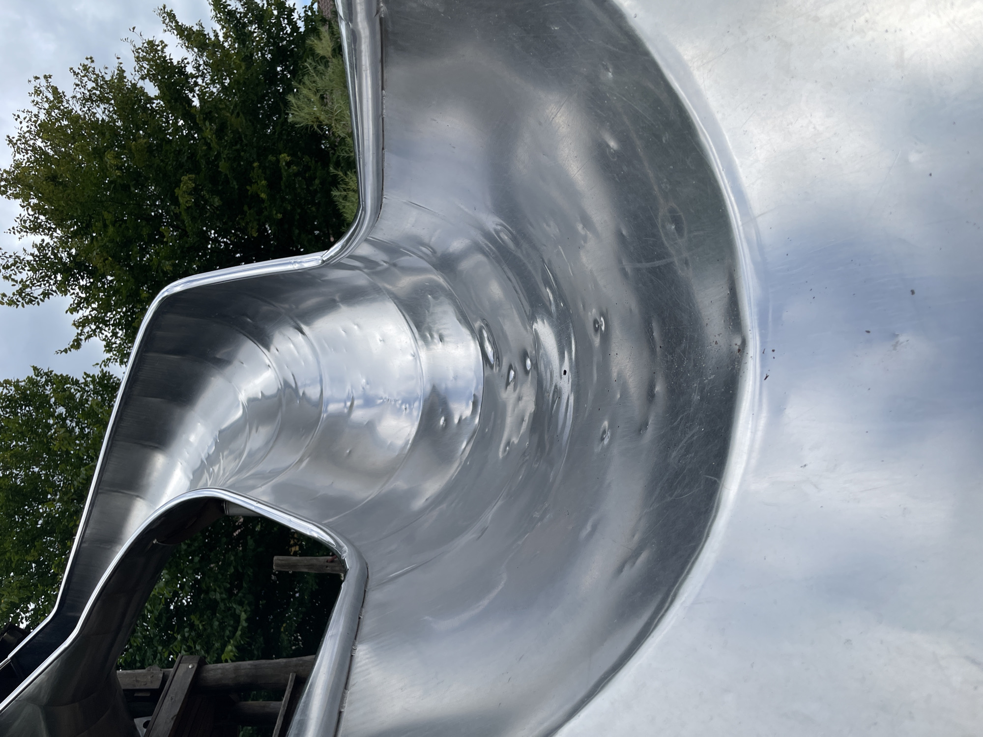 The image shows a close-up view from inside a shiny metal playground slide. The slide has a curved, tubular shape. The surface of the slide reflects the sky and surrounding greenery, with some visible dents and scratches. In the background, out of focus, are trees and a cloudy sky. On the left side of the image, parts of a wooden playground structure are visible. There are no people or animals present in the image. This slide is located in the outdoor play area of the German Maritime Museum in Bremerhaven, Germany.