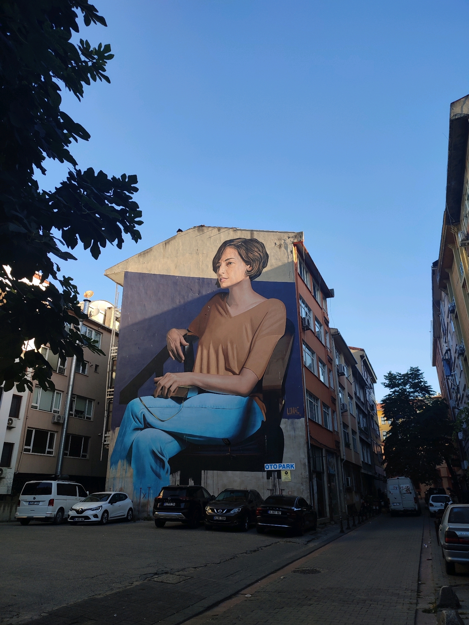 The image depicts a wide, paved street in Kadıköy, Turkey, flanked by multi-story buildings. On the left side of the street, a large mural of a woman sitting in a chair dominates the side of a building. The woman is depicted in a relaxed pose, wearing a brown shirt and blue pants. Her gaze is directed to the right. The mural is signed "LONAC". Below the mural, a white van and several cars are parked. The street is lined with trees on the right side, and more buildings with various colored facades. The sky is clear blue. The overall scene suggests an urban environment with a prominent piece of street art.