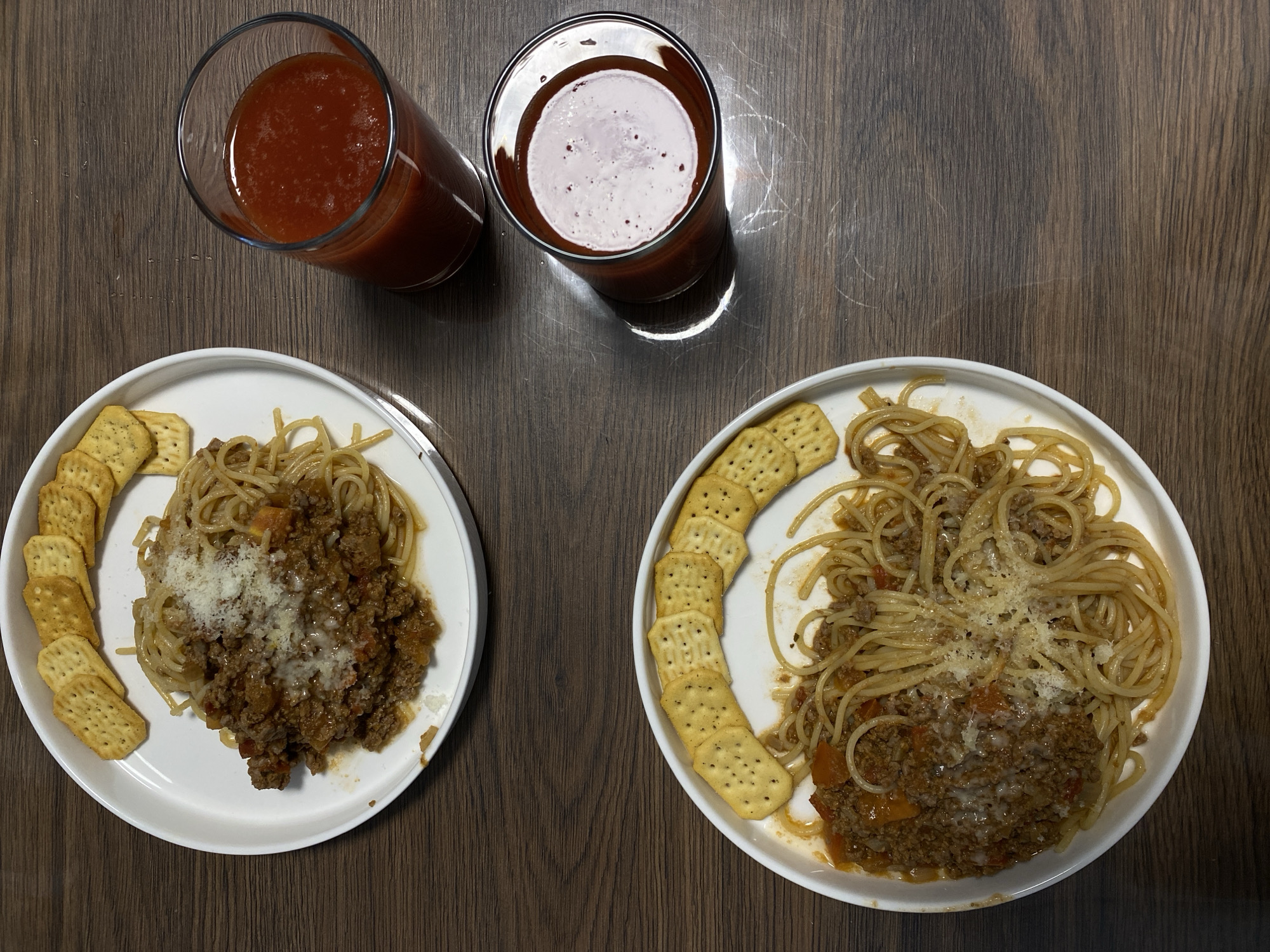The scene depicts an overhead view of a meal for two set on a dark brown wooden tabletop with a discernible grain pattern. Two identical white, round plates are positioned diagonally, with the top plate in the upper left quadrant and the bottom plate in the lower right quadrant. Each plate contains a serving of spaghetti coated in a dark reddish-brown meat sauce, consistent with bolognese, centrally adorned with a generous sprinkle of white, grated cheese. Along the outer edge of each plate, arranged in a semi-circular fashion, are approximately eight rectangular crackers, each bearing multiple small perforations. To the right of the upper plate and slightly above the lower plate are two clear glass tumblers, both filled with a homogeneous, opaque, reddish-orange liquid, characteristic of tomato juice. The surface of the liquid in the lower glass exhibits a lighter, frothy layer. No individuals or active interactions are visible within the frame. The scene is situated in the city of Saint Petersburg, Russia.