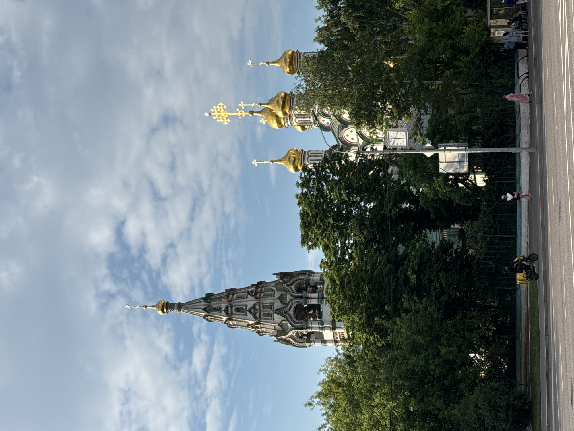 The image shows a view of the Church of St. Nicholas the Wonderworker in Khamovniki, Moscow, Russia. The church has a tall bell tower on the left and several golden onion domes on the right. A dense green canopy of trees partially obscures the view of the church. In the foreground, a street with traffic is visible. A delivery person on a scooter with a yellow insulated box is riding on the sidewalk. Further down the sidewalk, several people are standing and sitting, some at a bus stop. A clock is mounted on a pole near the church. The sky is blue with scattered clouds.