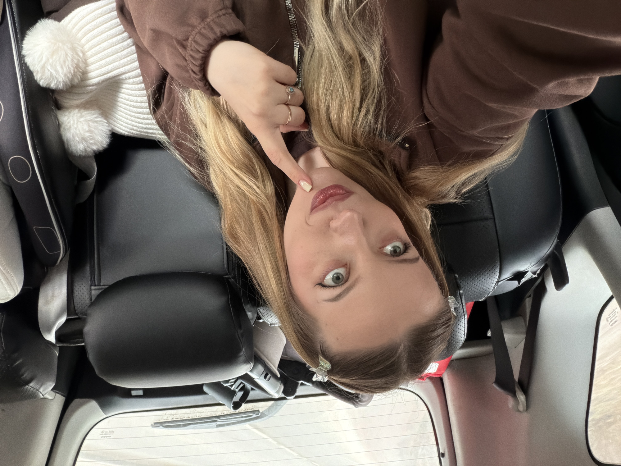 A woman with blonde hair and light skin is taking a selfie in the back seat of a vehicle. She is wearing a brown jacket over a white sweater and has rings on her fingers. She is pointing to her face. The interior of the car is visible, including black leather seats, a car seat, and a headrest. Outside the rear window is a view of what appears to be a blurred landscape, possibly indicating movement. The location is unknown, but the request indicates Вельмогово, Russia.