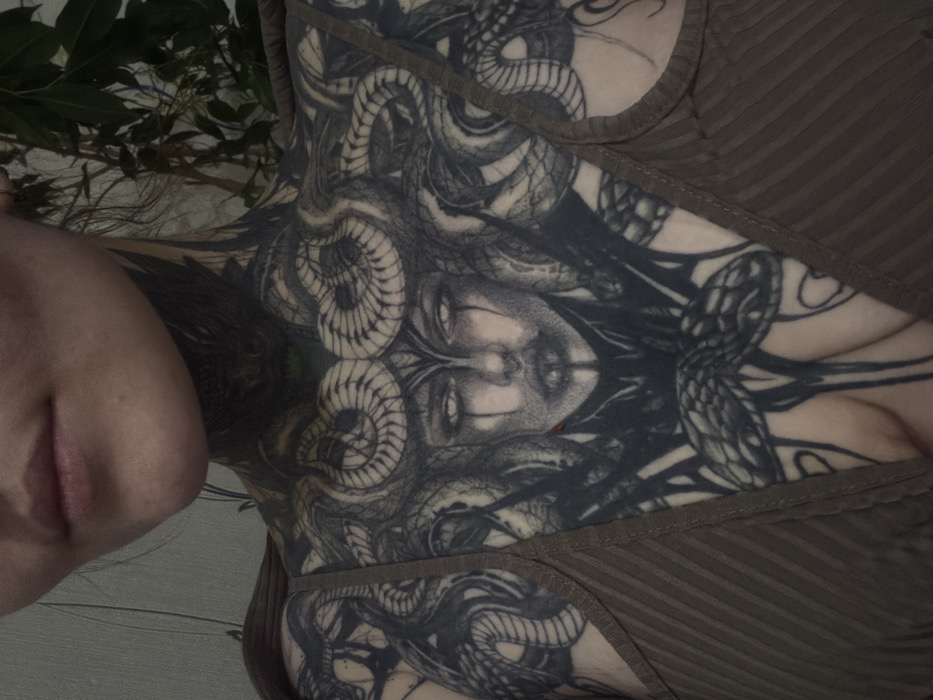 An image shows a woman's torso and part of her face. She's wearing a textured, olive-green top. A large, intricate Medusa tattoo covers her chest, featuring snakes forming hair around a symmetrical face. The composition has a slightly blurred quality and is rotated to the left. Green foliage is visible in the background.