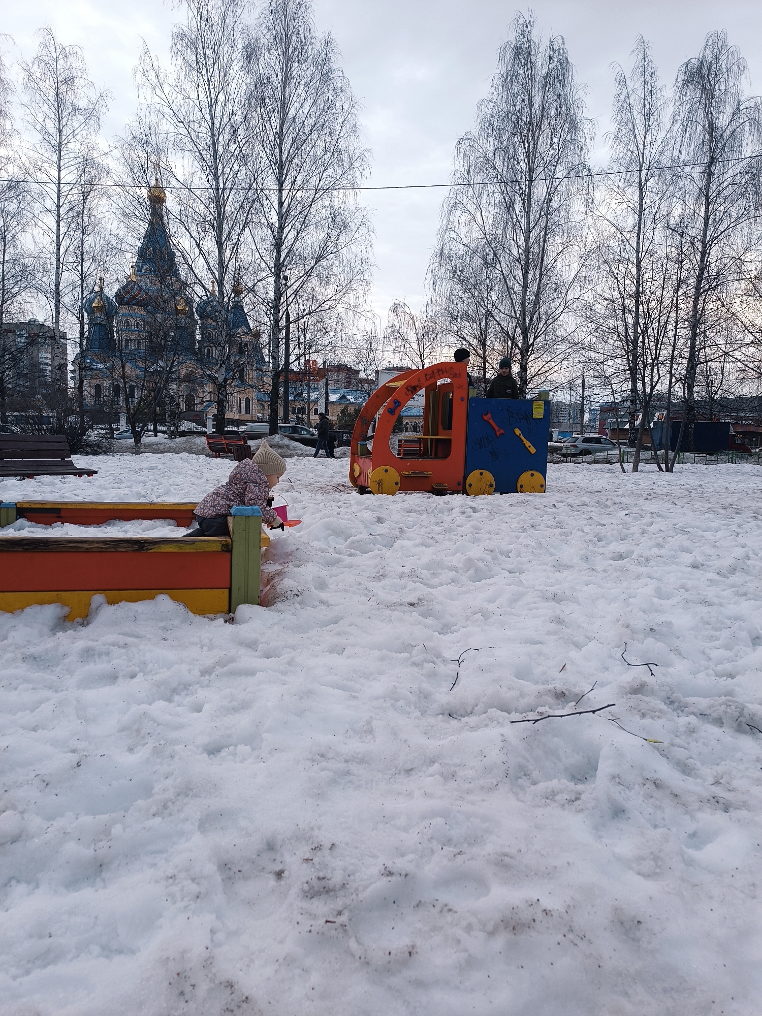 The image captures a snow-covered playground in Izhevsk, Russia. A young child, wearing a hat and jacket, plays in a sandbox. Nearby, a colorful play structure resembling a vehicle is occupied by two individuals, possibly children or young adults. In the background, a Russian Orthodox church with multiple domes is visible among bare trees, suggesting a winter setting. Other bare trees are scattered throughout the park area. The ground is entirely covered in snow. In the distance, buildings and cars indicate the location is within the city of Izhevsk.