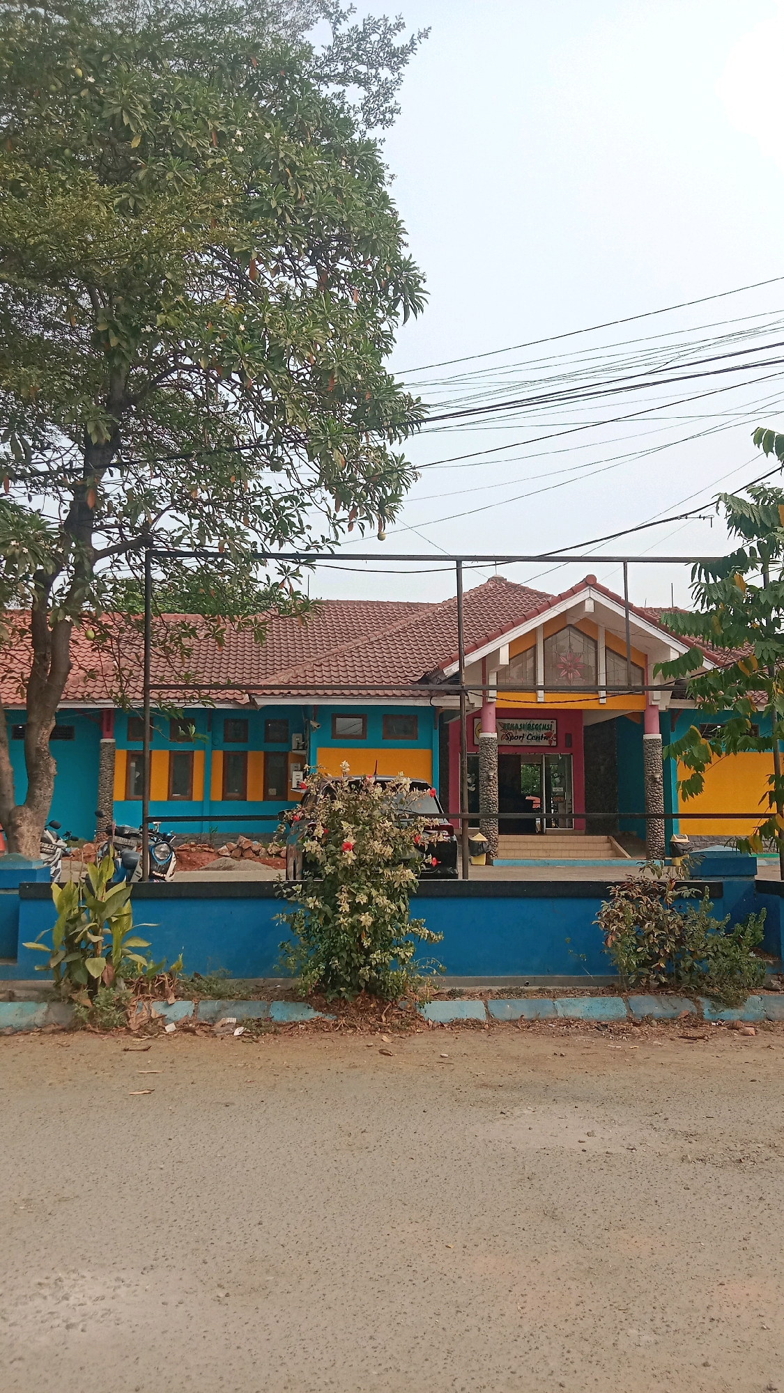 The image shows the front of a sport center in Wanasari, Indonesia. The building has a red tiled roof and is painted in vibrant colors, including teal on the main structure, with accents of yellow and orange on the window frames and the main entrance. The entrance features a pointed gable with decorative glass. A sign above the entrance reads "FASILITAS KESEHATAN Sport Centre." Several columns support the roof over the entrance area, painted in pink and grey stone patterns. A black car is parked in front of the building, and several motorcycles are visible to the left. The foreground shows a paved road and a blue concrete curb with some vegetation and debris. Overhead, power lines are strung across the sky. The overall scene depicts a public facility in a tropical or sub-tropical climate, with clear daylight conditions and a bright, overcast sky.
