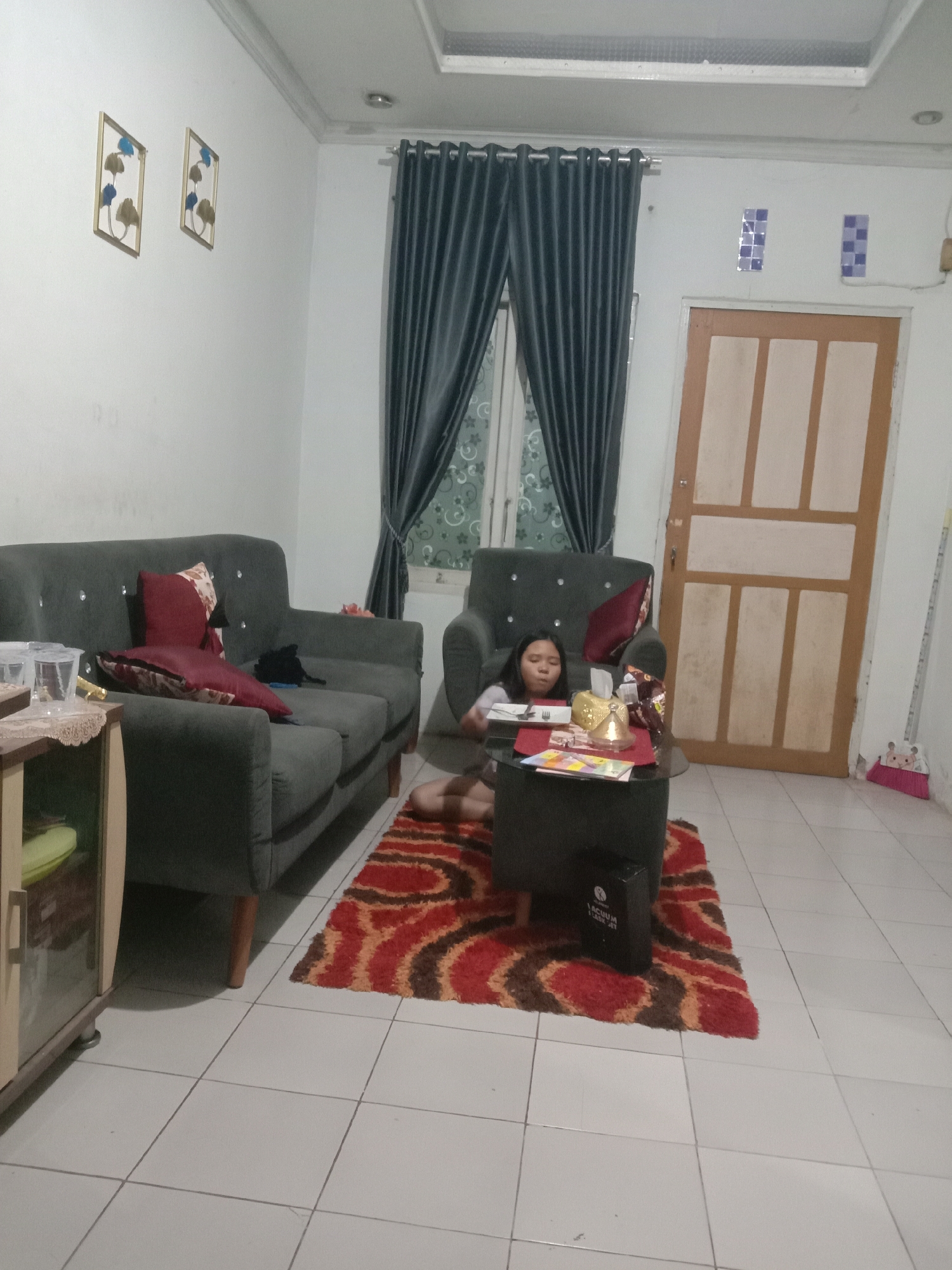 This is a medium shot, eye-level, indoor scene of a living room in a home in Kwandang, Indonesia. The room is sparsely furnished with a dark grey sofa on the left, a single armchair on the right, a small round coffee table between them, and a large rug with a red, orange, and brown patterned design in the center. The walls are white, and the floor is tiled with light grey squares.

In the armchair, a young girl with dark hair is seated and appears to be looking at something on the coffee table. She is wearing a light-colored shirt. On the coffee table, there are several items including a tissue box, what appear to be some books or magazines, and a plate with cutlery. A dark object with writing on it, possibly a bag or a case, is placed on the rug near the coffee table.

To the left of the sofa is a small cabinet with a glass door, partially obscuring some items inside. A dark grey curtain hangs behind the sofa, and another curtain, possibly with a patterned design, is visible behind the armchair. A door with a light wood frame and multiple rectangular panels is situated to the right of the armchair. On the wall above the door, there are two small decorative elements consisting of blue and silver squares. To the far right of the image, a broom with a pink dustpan is leaning against the wall. Two framed decorative pictures, each containing abstract blue and gold leaf shapes, are mounted on the wall above the sofa. The ceiling has recessed lighting fixtures.