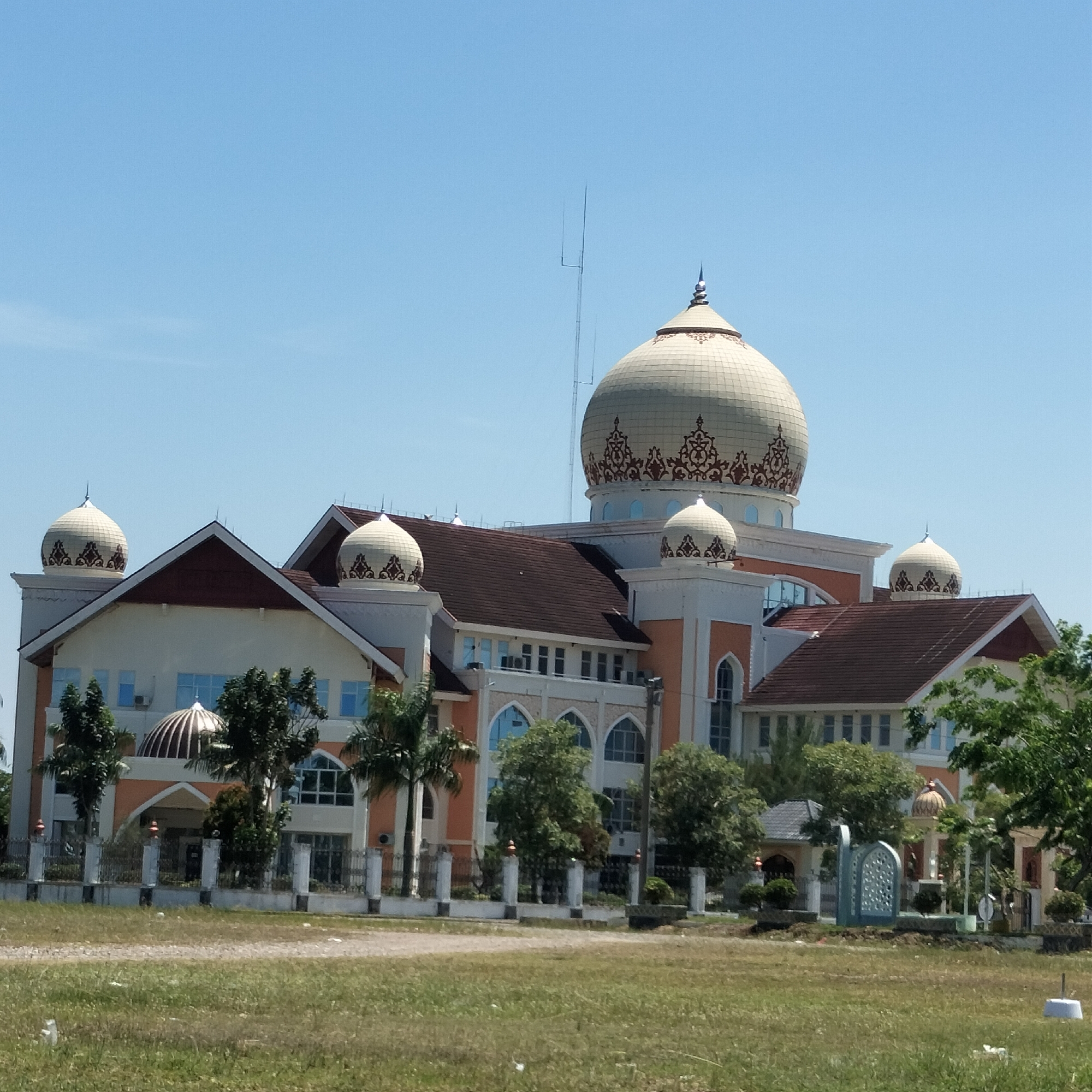 The image shows a large, ornate building, likely a mosque or government building, in Meunasahaluedrienlhoksukon Barat, Indonesia. The building has a central, large dome with decorative patterns, and several smaller domes on the rooftops of the surrounding structures. The architecture features arches, windows, and a color scheme of white, peach, and brown. A tall antenna is visible behind the main dome. The building is surrounded by a grassy field and a low fence, with several trees and landscaping features nearby. The sky is clear and blue.