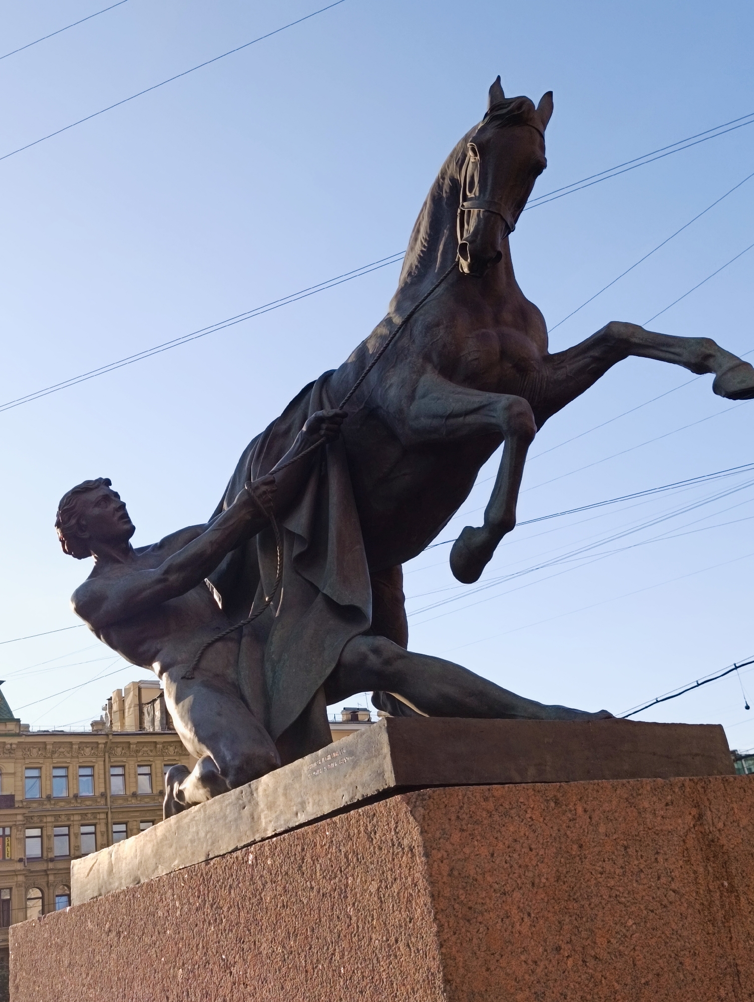 The image shows a bronze statue depicting a man struggling to control a rearing horse. The man is on his knees, pulling on a rope that is wrapped around the horse's neck and draped over its body. The horse is on its hind legs, its forelegs in the air, and its head turned away. The statue is positioned on a granite pedestal, which is mounted on a larger, rough-hewn granite base. In the background, a building with classical architectural elements, including ornate facades and rows of windows, can be seen. The sky is a clear, light blue with several power lines crisscrossing it. This is a detail of the Monument to the Horse Tamers by Petr Klodt, located on the Anichkov Bridge over the Fontanka River in Saint Petersburg, Russia.