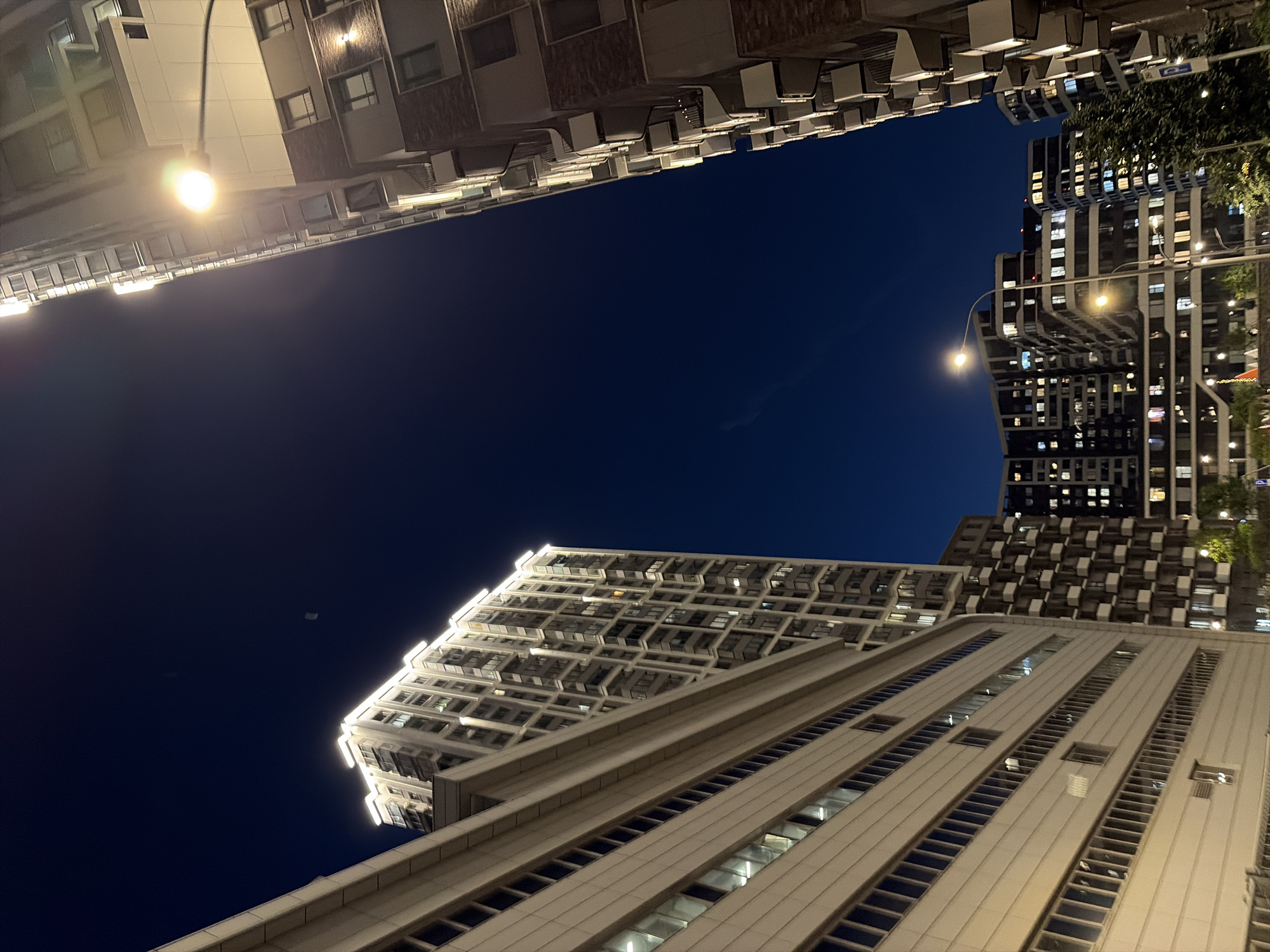 The image depicts a night view of a modern urban landscape in Moscow, Russia. Multiple high-rise buildings are visible, with many windows illuminated, indicating occupancy. The architecture appears to be contemporary, with some buildings featuring balconies and distinct facade designs. A streetlamp with a bright light source is prominently featured in the foreground, casting a glow on the surrounding structures. The sky is a deep, dark blue, characteristic of nighttime. The perspective is from ground level, looking upwards at the buildings. No people are clearly visible in the scene. The overall impression is one of a well-lit, residential or mixed-use urban environment at night.
