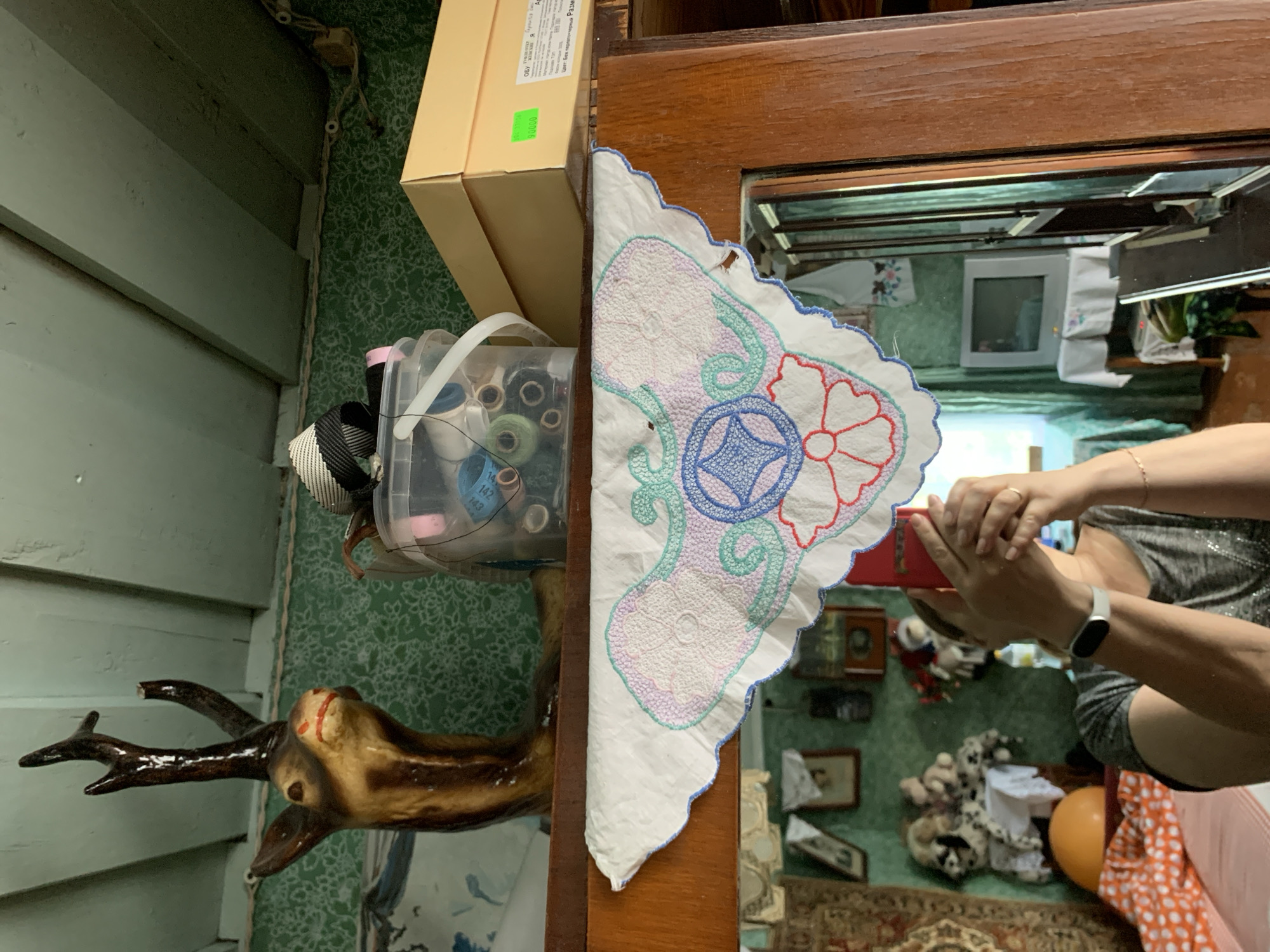 This image captures an interior scene in Відзаўскі сельскі Савет, Belarus, primarily showing a shelf and a mirror reflection.
On the uppermost shelf, a mounted deer head sculpture, possibly a replica, is visible on the left, featuring dark brown antlers with one tip broken off, a light brown face, and red painted nostrils. To its right, a clear plastic bucket holds numerous spools of thread and other sewing accessories, including a white measuring tape. Further right, stacked beige cardboard shoeboxes are present, one displaying a green price sticker with Russian text and details about footwear ("ОБУ", "Я", "90000", "Цвет: Беж перламутровый размер"). The background wall is covered with a green wallpaper featuring a faint white floral pattern. Above this, light green wooden ceiling beams are visible, with exposed electrical wiring running along the wall-ceiling joint.
Below the top shelf, a wooden surface supports a white embroidered doily with a scalloped edge. The embroidery features a central blue star-like motif, surrounded by light purple, green, and red floral and swirling patterns.
The lower half of the image is dominated by the reflection in a large mirror, framed by dark brown wood on the right. A person is visible in the reflection, holding a red smartphone with both hands to take the picture. The individual wears a grey short-sleeved top with subtle sparkle, a white fitness tracker on their left wrist, and a gold ring on the ring finger of their right hand. The reflected room also features the same green patterned wallpaper. On the left, a tapestry or rug hangs on the wall, and beneath it, a surface holds several stuffed animals or dolls. An orange domed object and orange-and-white patterned fabric are visible in the lower-left corner of the reflection, possibly on a bed or seating. In the center-right of the reflection, a white, older model CRT television sits on a light-colored stand covered with a white tablecloth. Light green curtains are visible through a window behind the television, and a plastic bottle stands nearby. The room appears somewhat cluttered with various household items.