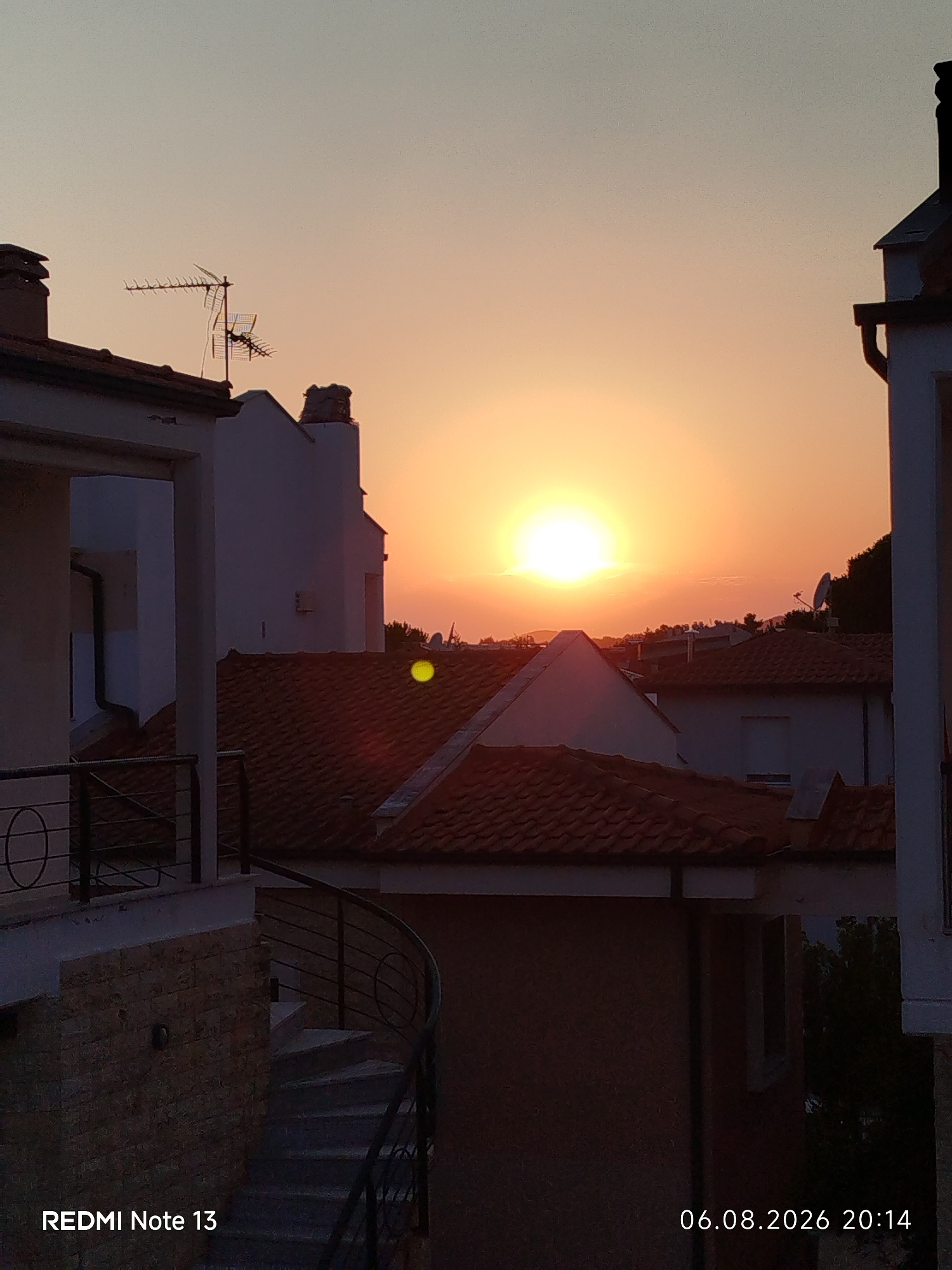 A vertical, low-angle shot captures a sunset over rooftops in Sithonia Municipal Unit, Greece. The sky is a vibrant gradient of orange and yellow, with the sun appearing as a bright, glowing orb near the center. Silhouetted against the sky are several buildings with tiled roofs and antennae. In the foreground, a stone-clad building with a spiral staircase and metal railings is visible, partially illuminated by the fading sunlight. A yellow circular lens flare is present in the mid-ground, slightly to the left of the sun. The bottom left corner of the image displays the text "REDMI Note 13" and the bottom right corner shows the date and time "06.08.2026 20:14".