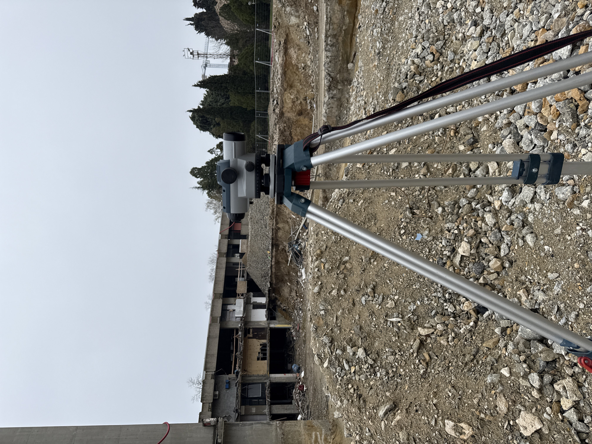 The image depicts a surveying instrument, likely a level, mounted on a tripod. The instrument is positioned in a construction or demolition zone. The foreground consists of a gravelly surface. In the background is a partially demolished building, revealing the building's interior, and trees are also visible. The sky is overcast. The presence of the surveying instrument suggests that site measurements are being taken, likely related to construction or demolition activities.
While I can't definitively confirm the location, the scene characteristics suggest an urban construction site. I lack the information to confirm that this is in Varna, Bulgaria.
